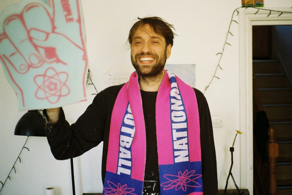 Photograph of a man from the waist up, wearing a pink and purple football scarf and holding up his right hand, on which he wears a giant foam hand with a pointed finger. 