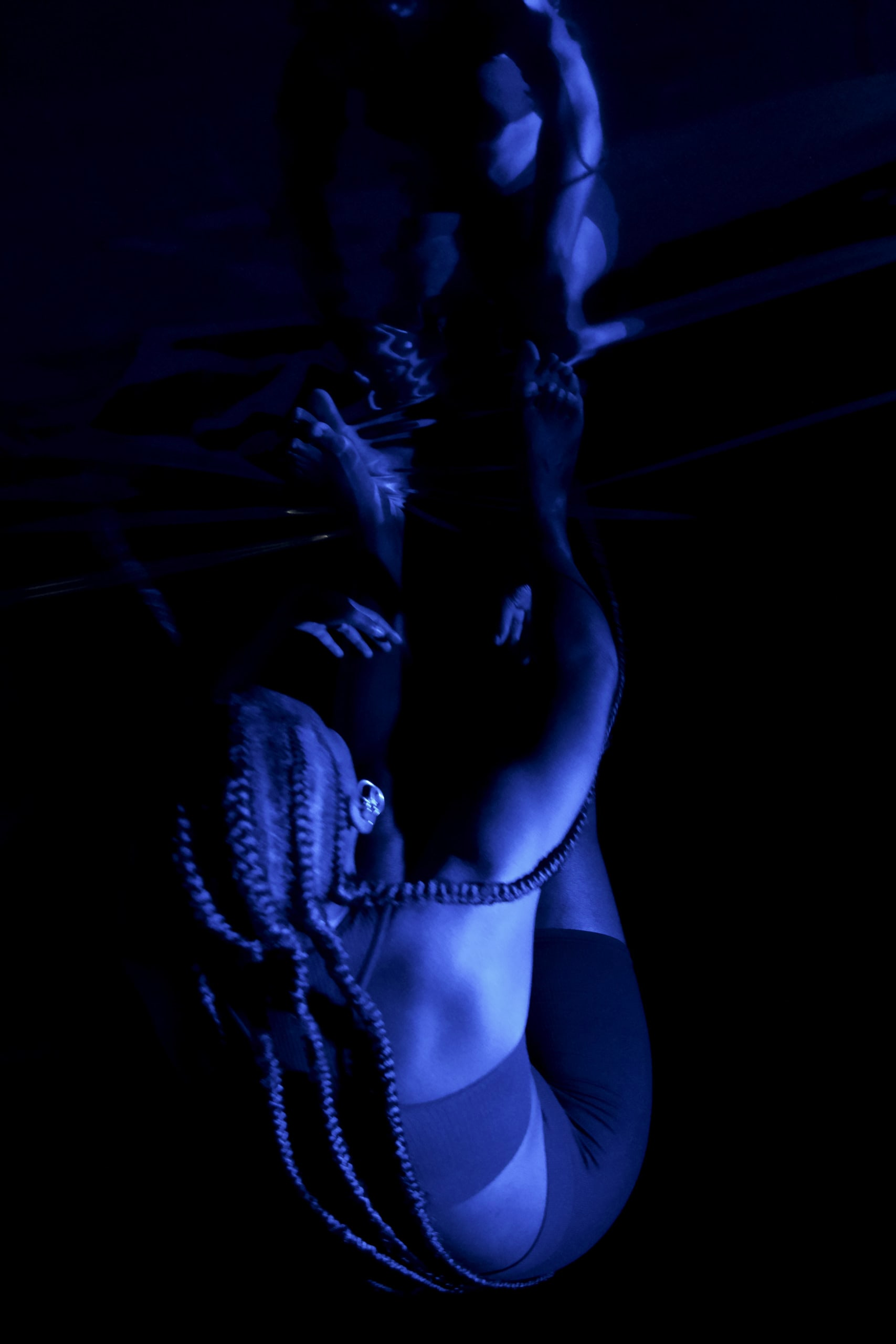 An blue tinted aerial image of woman bent over, reaching up to her legs, which are at the top of the image, on a black background.