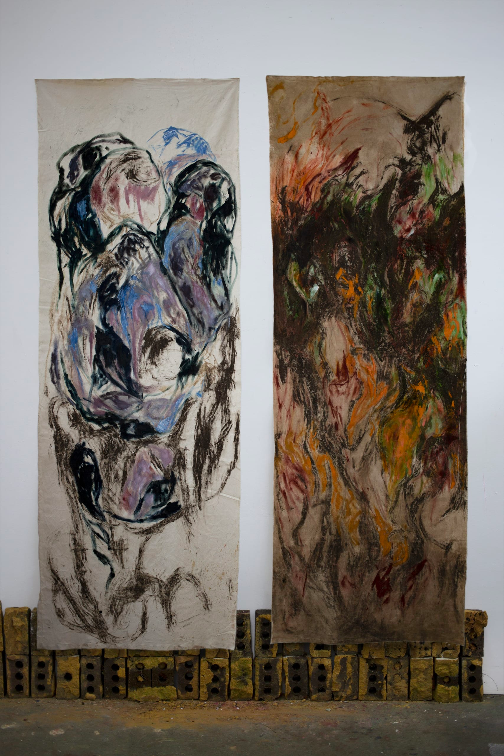 Photograph of two rectangular abstract paintings, on what looks like cloth, hanging on a wall, side by side, one in shades of blue, pink and black, the other in fiery orange, red, green and black.