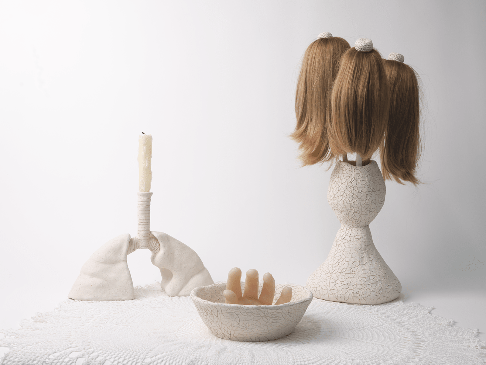 Image of a white candlestick, white bowl with a hand poking out of it, and a white vase containing three short poles with long brown hair attached, on a white round cloth on a white background.
