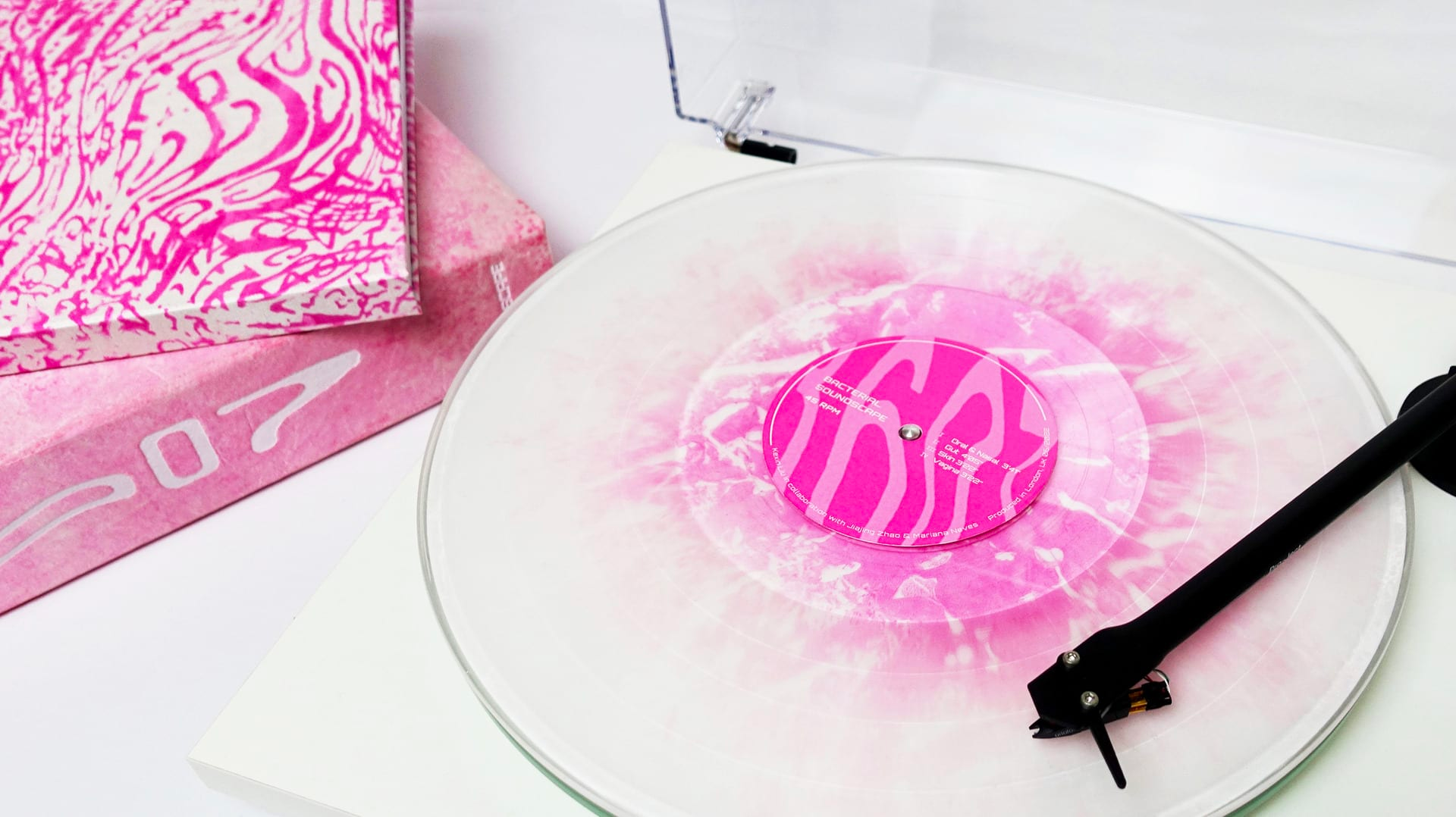 Close-up image of a white record player turntable with a black tone arm playing a pink and white or clear record, with the corners of two stacked pink and white shallow boxes next to it.