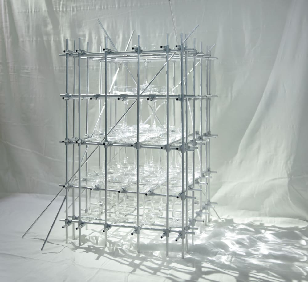 Photograph of a metal scaffolding-like structure, creating a grid, on which are placed lots of glass objects, against a white backdrop.