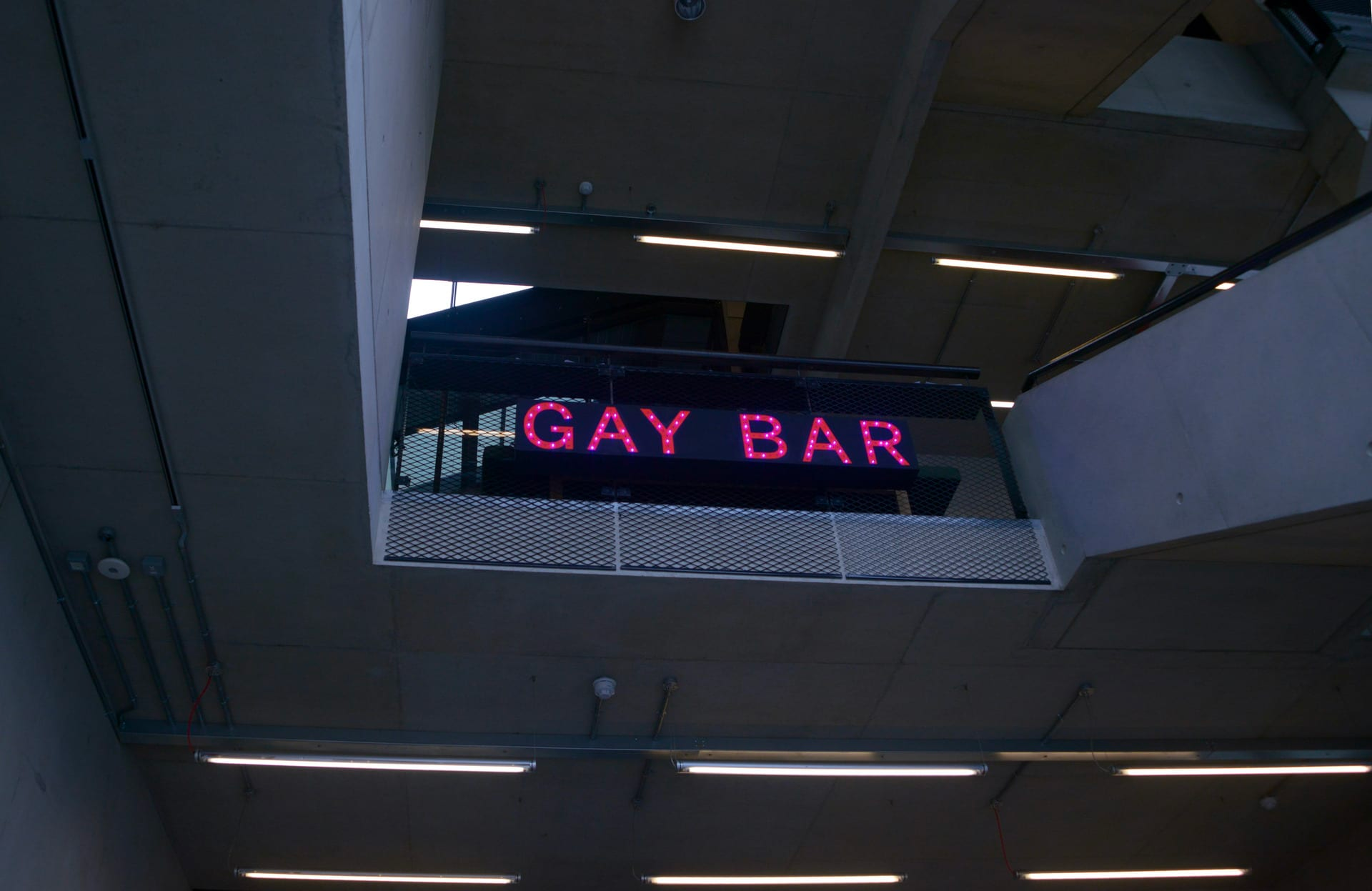Photograph looking upwards to a black, interior balcony with pink illuminated letters spelling ‘Gay Bar’.