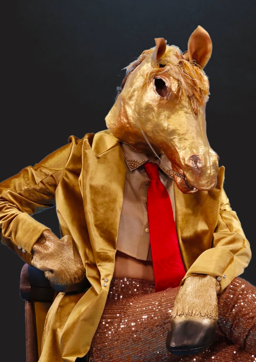 Photograph portrait of a person posed on a chair in a shiny gold jacket and sequinned trousers wearing a golden horse’s head mask and hooves.