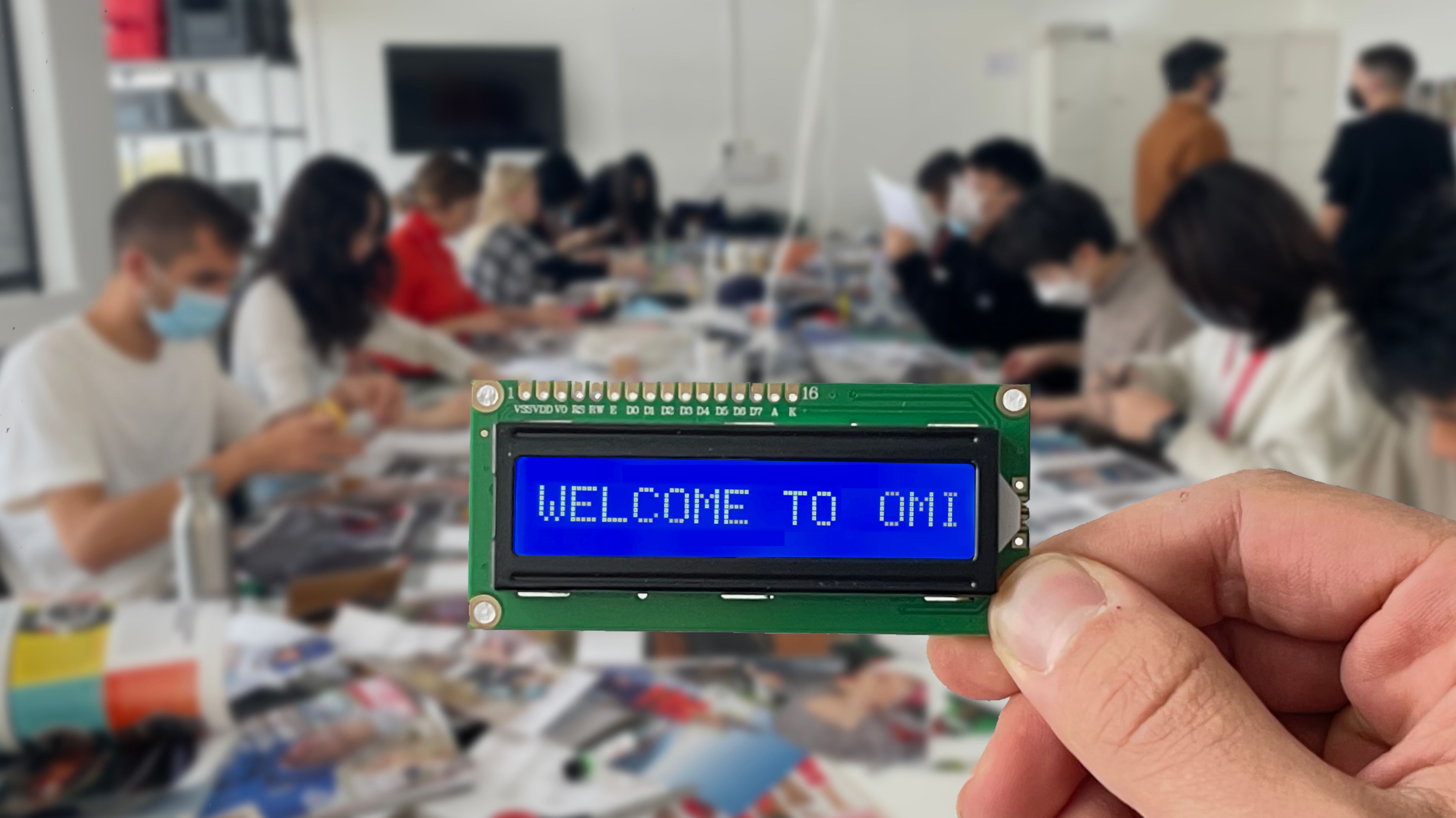 Photograph of a hand holding up a computer chip with the words 'Welcome to OMI' on it, in front of a long table covered in materials, at which people are sat, working, on either side.