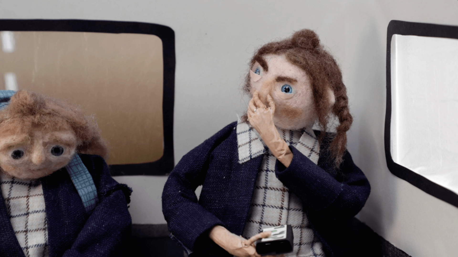 Photograph of two model figures – one holding her nose - made from felted wool. The figures are wearing the same uniform and sat on what looks like the back corner of a bus.