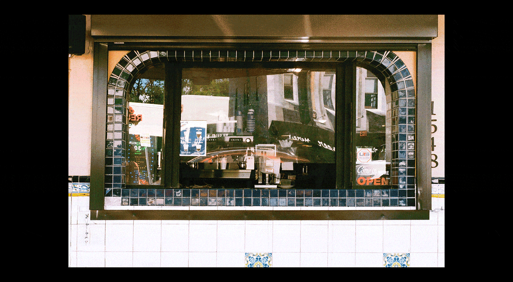 Photograph of what appears to be a rectangular window, on a partially tiled wall, which looks into to a café and is reflecting other nearby buildings.