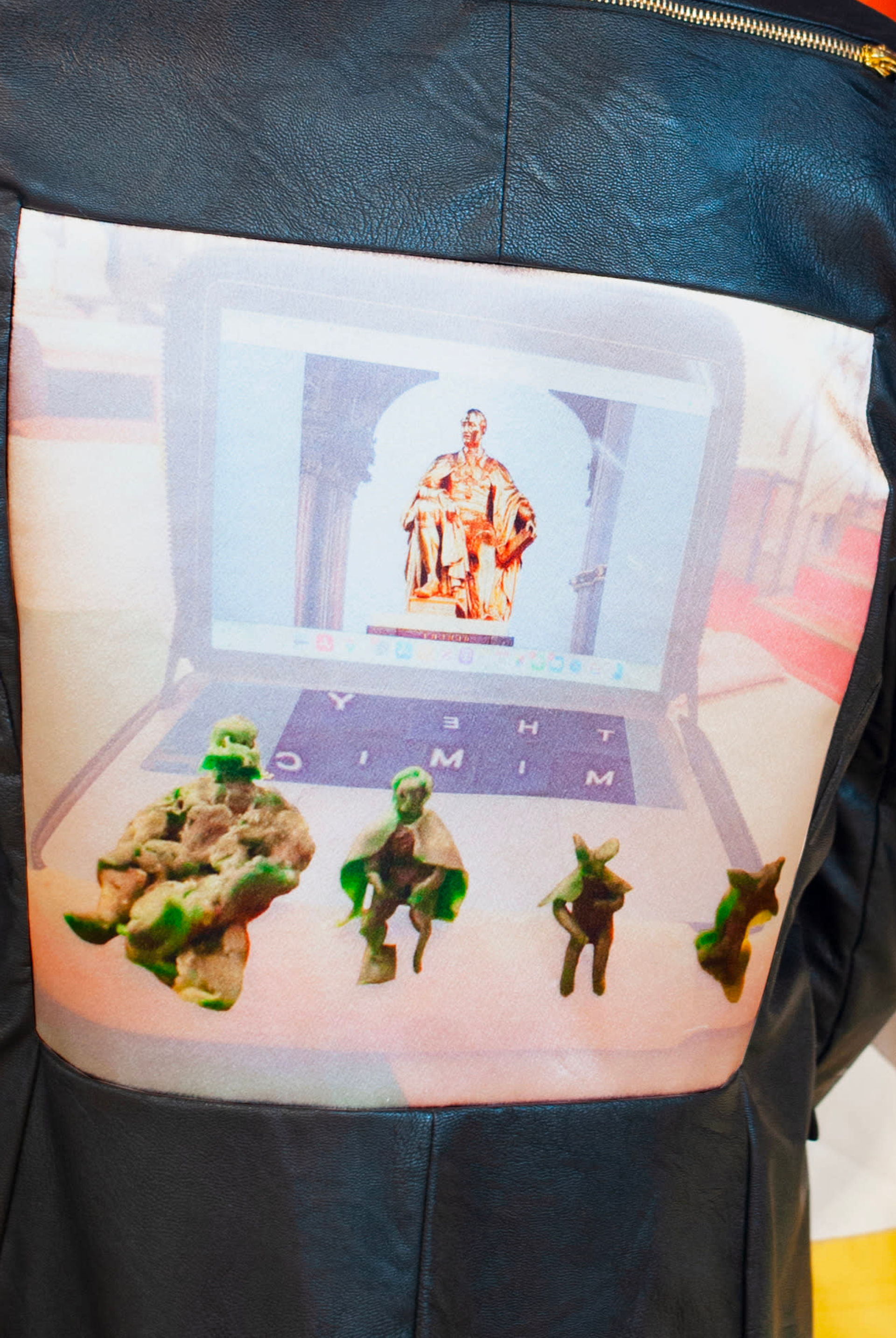 Image of, what looks like, a close-up on the back of someone wearing a black leather jacket. There is an image attached to the jacket, featuring green figures and a seated golden figure. By Franziska Windolf