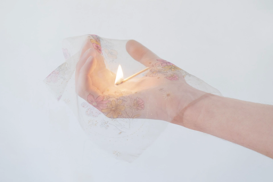 A photograph of an upturned hand, covered by a square of sheer fabric, with a lit match lying on top.
