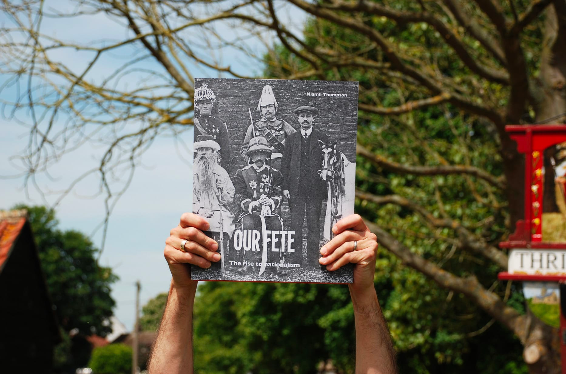 Photograph of a pair of hands, in an outdoor setting, holding up a book towards the camera, which has part of a black and white photographic group portrait on the cover, and the title ‘Our Fete’.