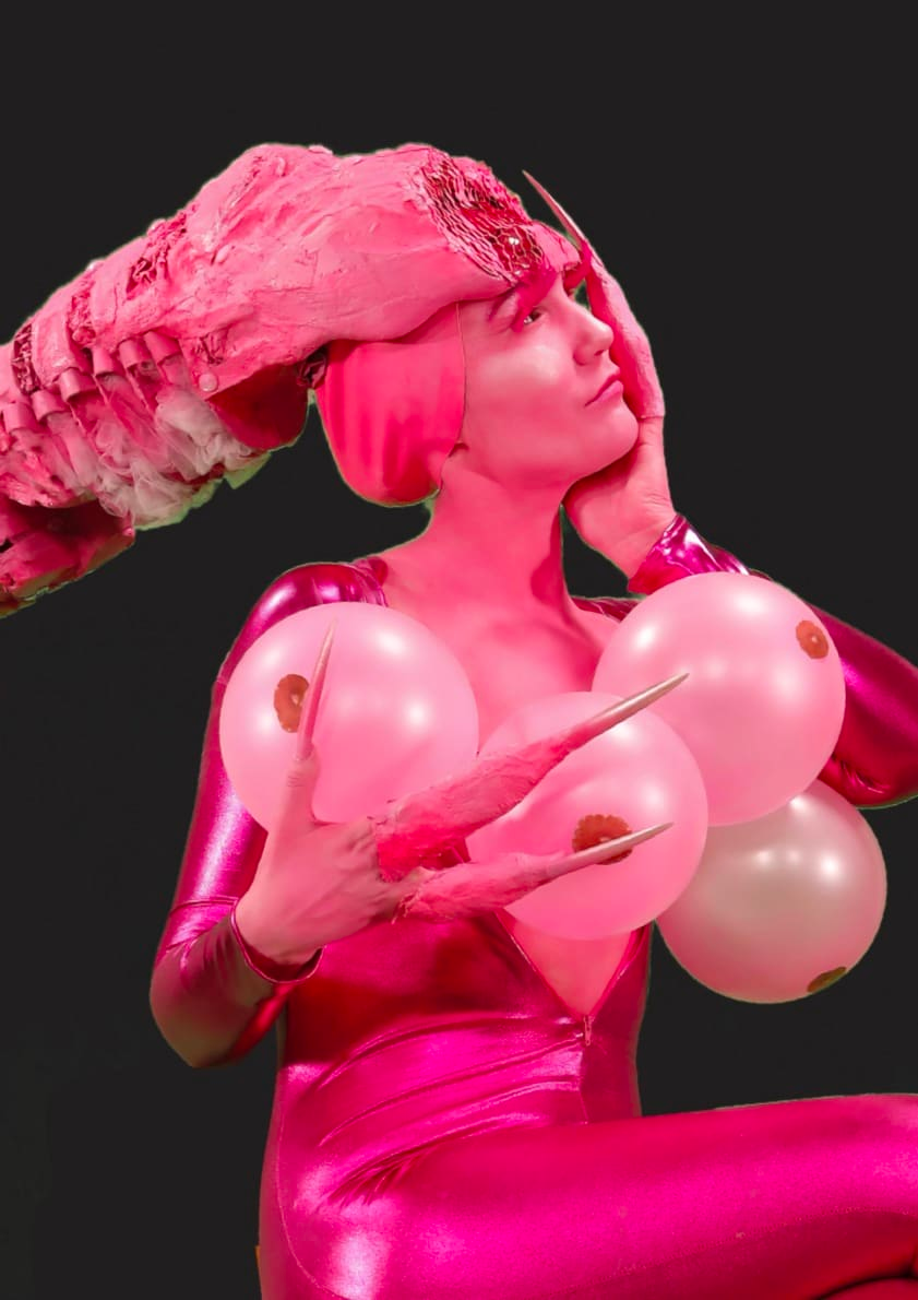 Photograph of a person in pink light wearing a tight pink bodysuit and a large pink headdress. She holds pink balloons with what looks like nipples on them, in her hands with three long pointed claws.