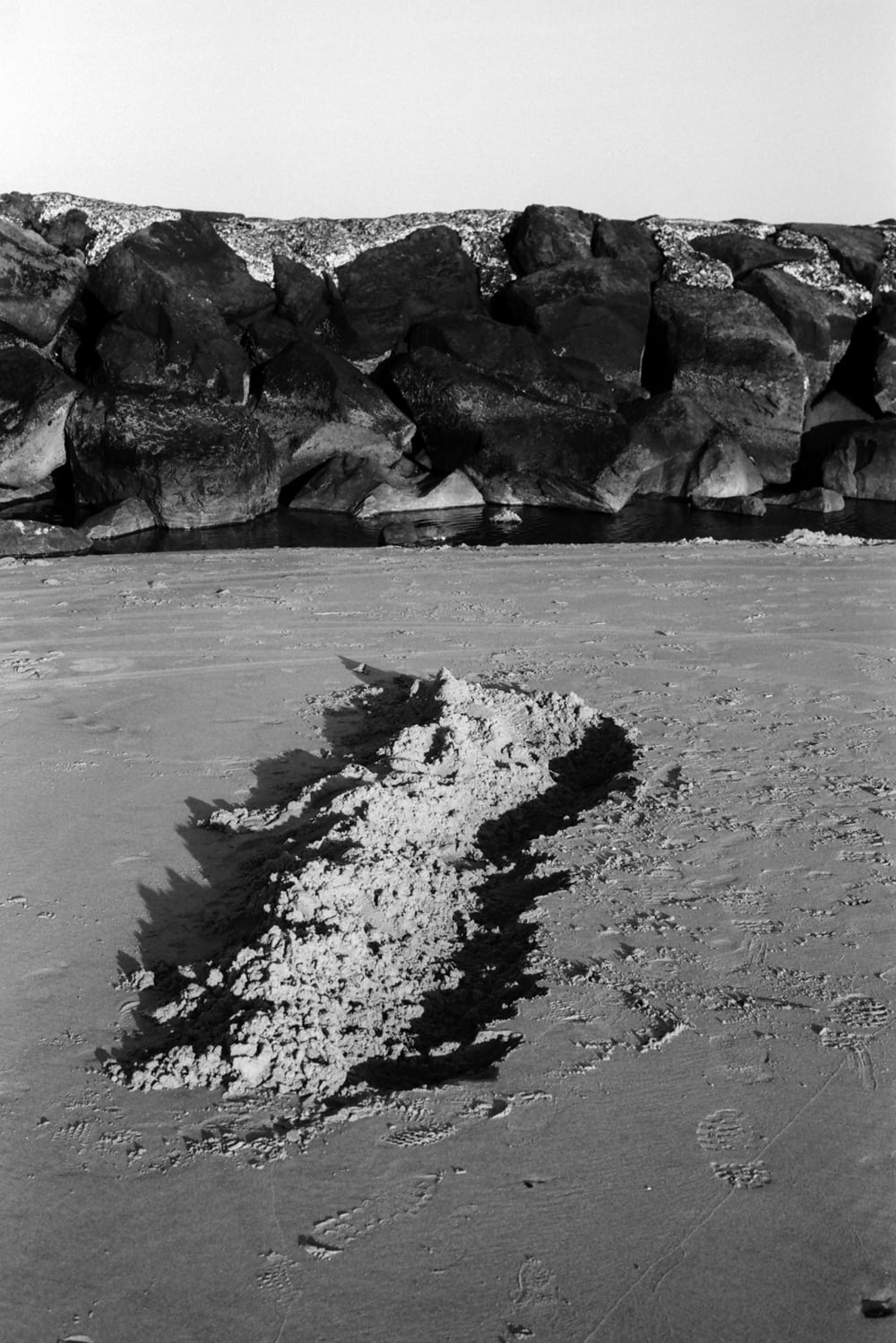 Black and white image of what looks like a length of shallow trench dug into sand, with footprints made by boots alongside, on what appears to be a beach with water and rocks in the background.