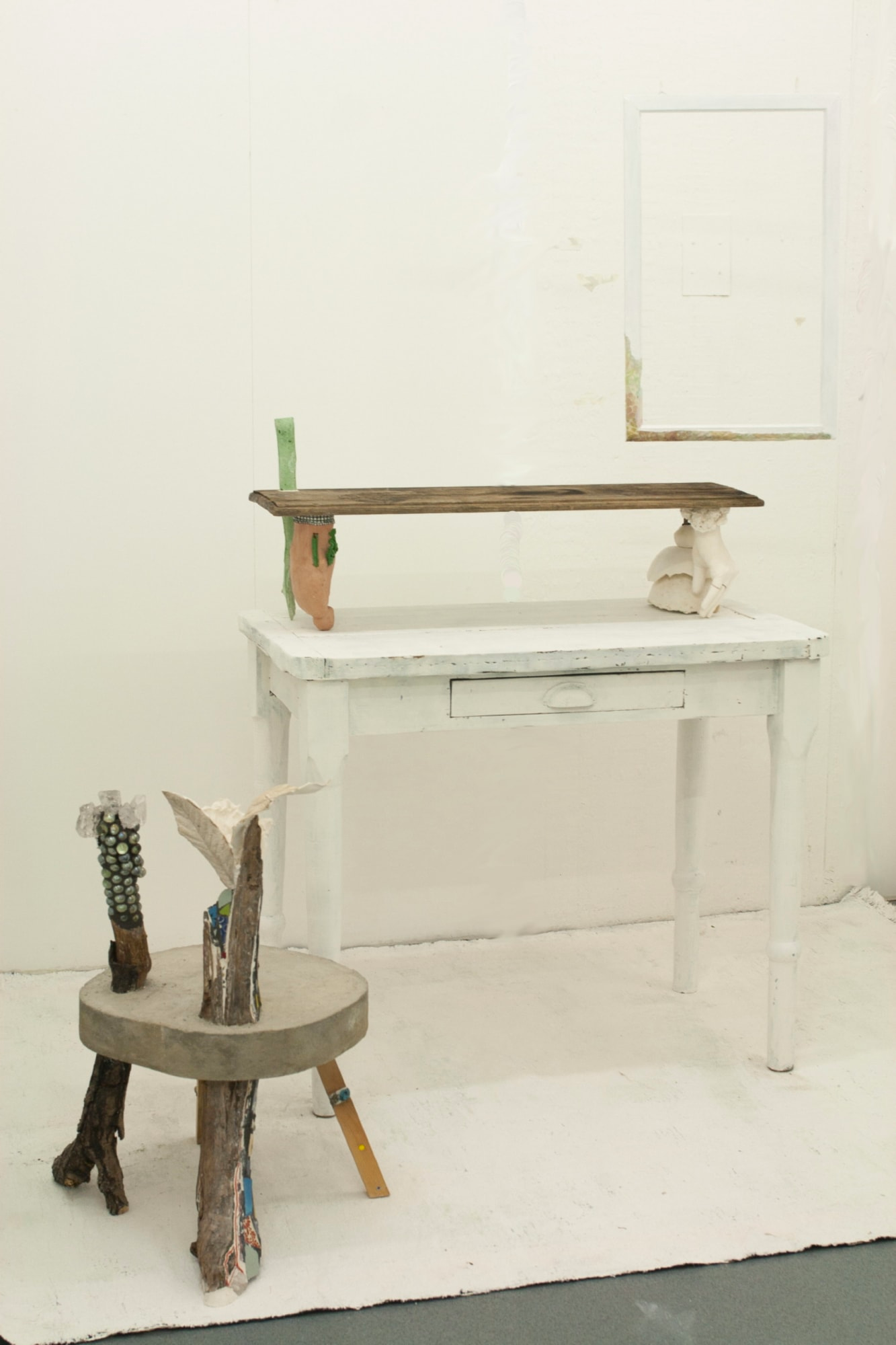 Image of a wooden table with one drawer, painted white, with a sort of shelf on it, alongside a wooden stool made from various items, against a white wall.