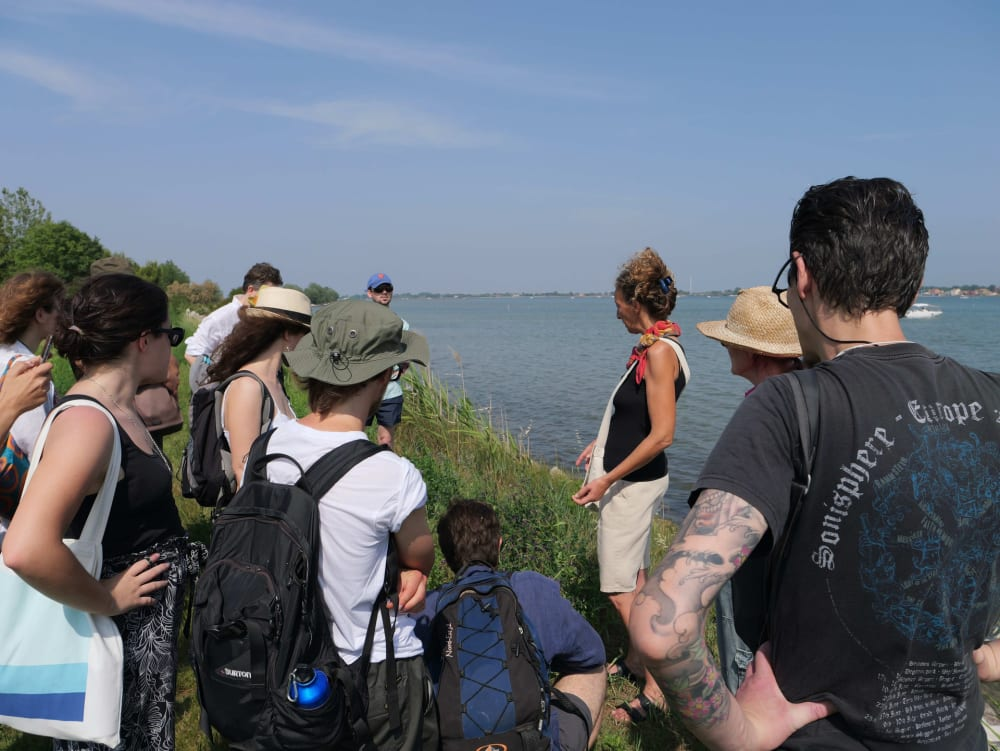 Photograph of a group of people, some with their backs to the camera, gathered around a woman who appears to be addressing them, on a grassy area next to a large body of water.