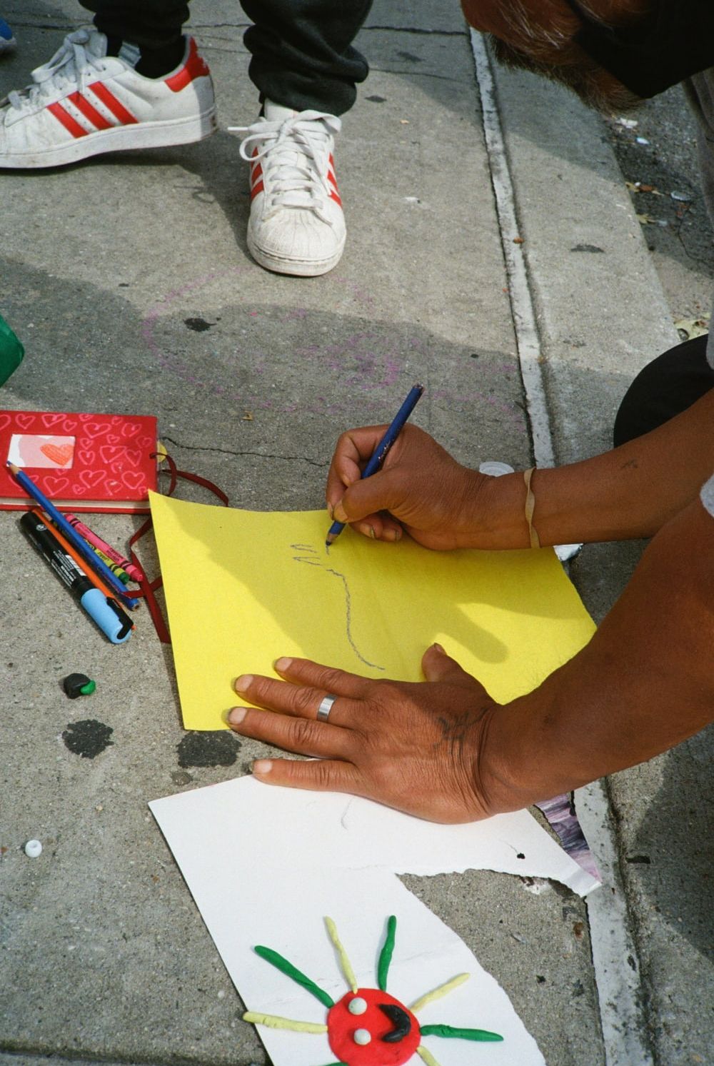 Photograph of a pair of hands drawing on yellow paper placed on the concrete ground. Next to the paper are pens, a plasticine image of the sun and another person’s feet wearing trainers.