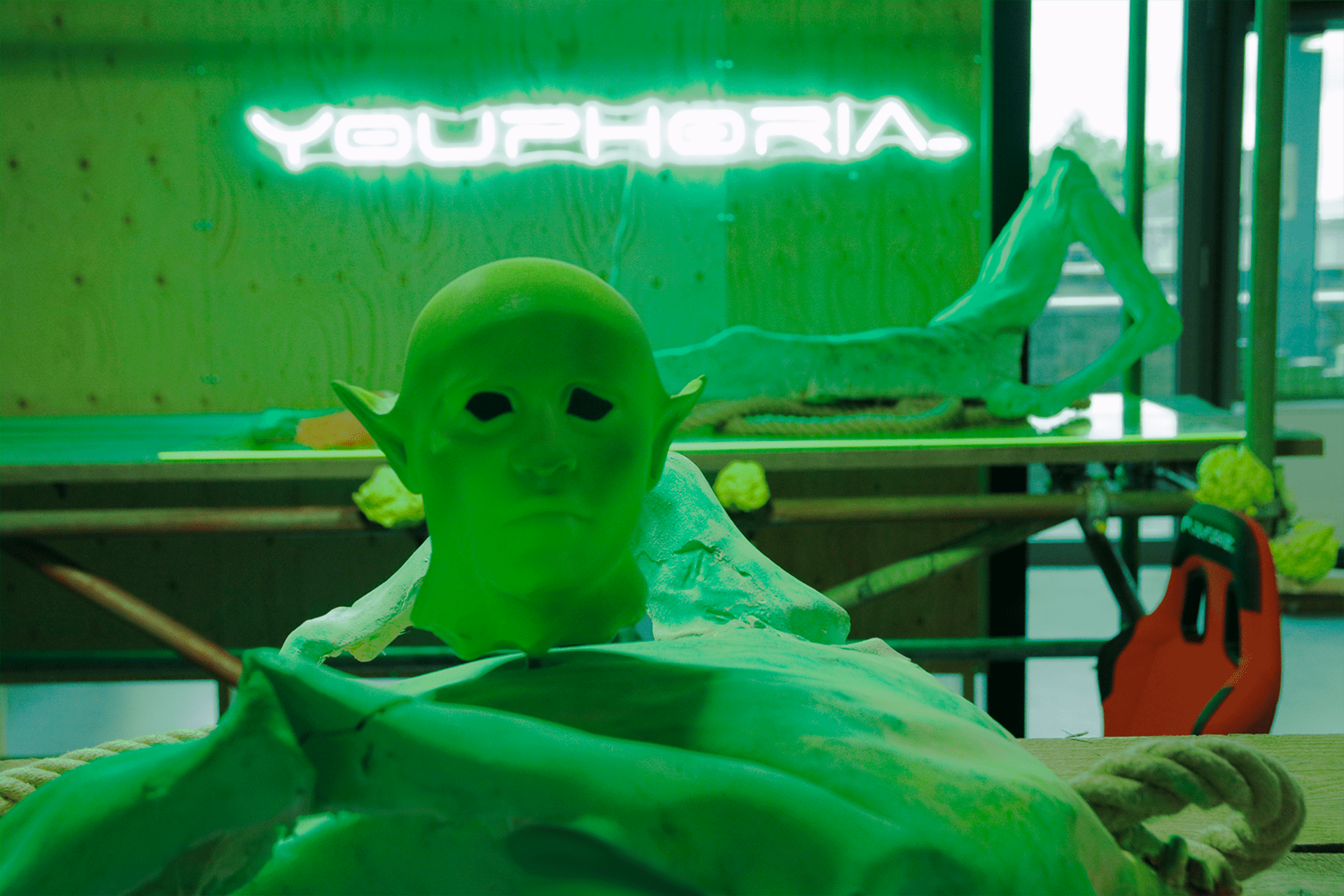 A photograph of a life-size mask of a head with large, pointed ears, and a semi-reclined headless figure on a table. A neon sign in the background, which reads ‘youphoria’, casts a green light over the objects.
