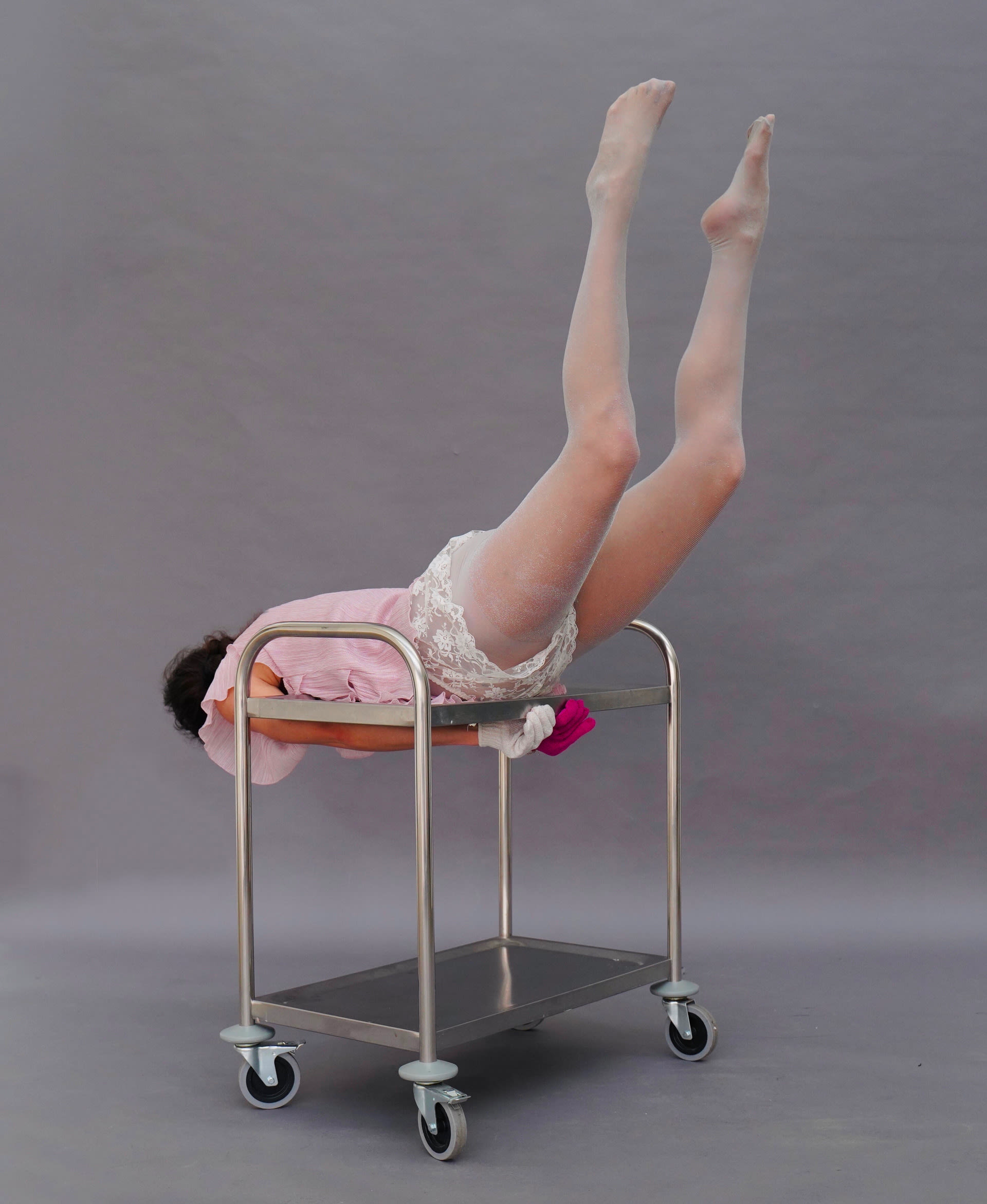 Image of a person turned to the side and away from the camera, face down on top of a silver food trolley with wheels, with their legs lifted up into the air, on a grey background.