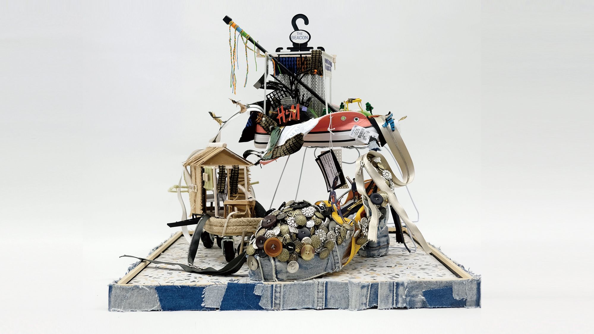 A pyramid of random objects including buttons, a split open trainer, sandal and topped with a plastic coat hanger, on a square slightly raised platform, edged with scraps of denim.