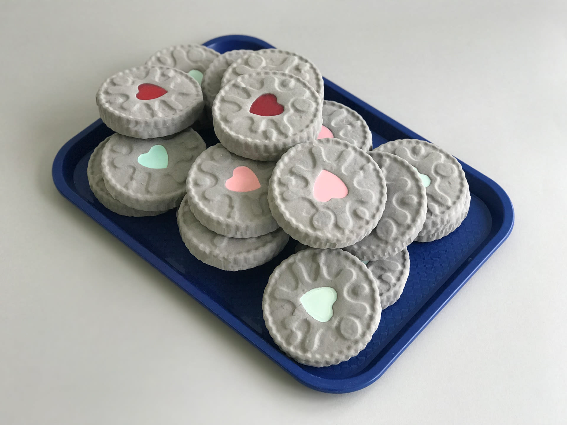 Photograph of a blue rectangular tray filled with biscuits resembling Jammy Dodger made from a hard grey substance, with heart-shaped centres filled in either pale green, pale pink or red.