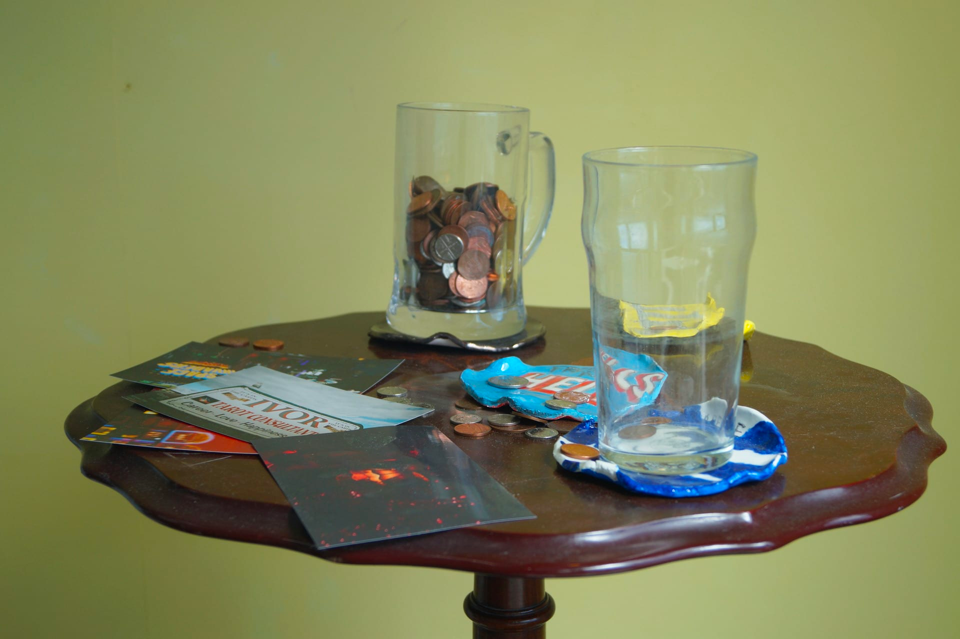 Photograph close-up of a dark wood pedestal table, with coins, what looks like flyers and three coloured discs scattered on it and two pint glasses, one empty and one filled with coins.