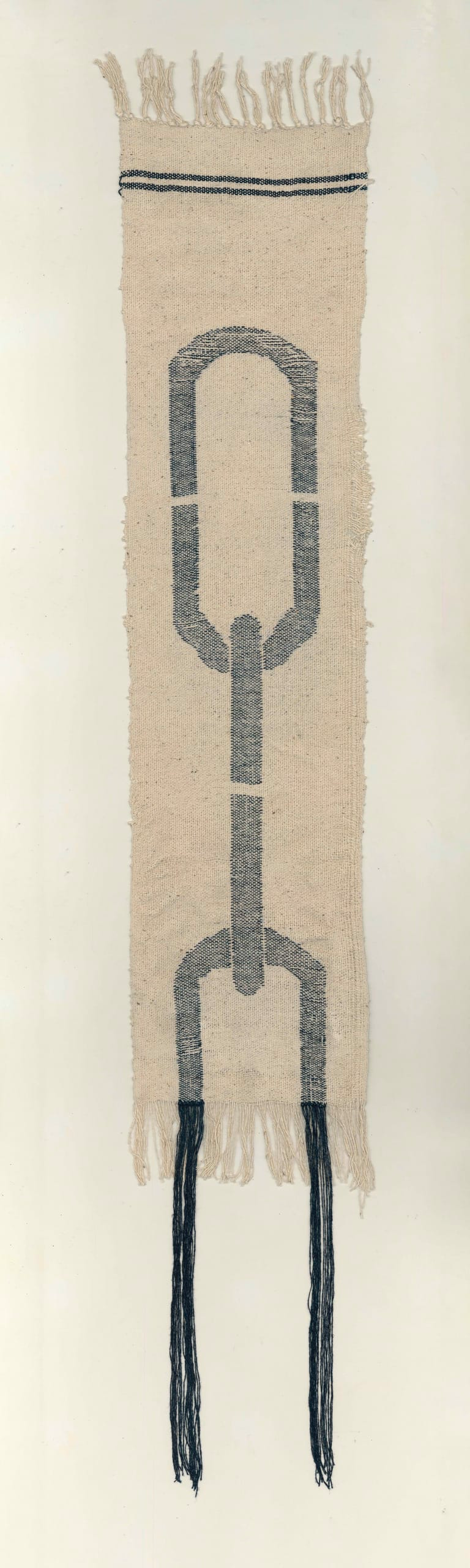 A photograph of a beige knitted or woven length of tasselled fabric, featuring a large black chain-like design, with one link open at the edge of the fabric, its ends trailing as two long tassels. 