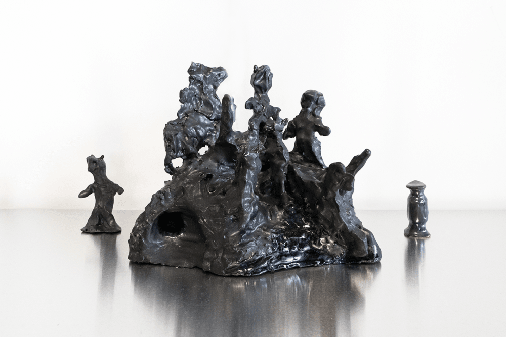 Image of an almost black sculpture, of figures emerging from a rocky base and another figure in the same style stood to the left of it and a small shiny black object to the right.
