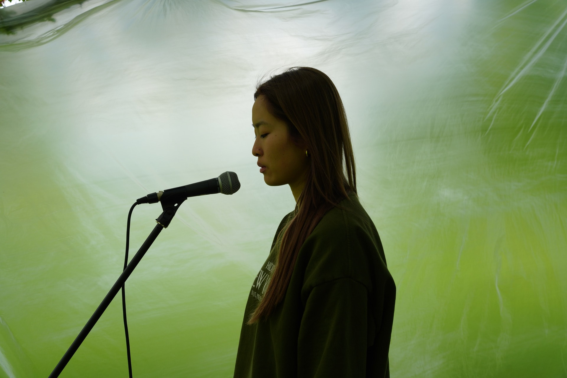 Photograph of a woman from the waist up, in profile, with her eyes closed, stood in front of a microphone on a stand, against a green background.