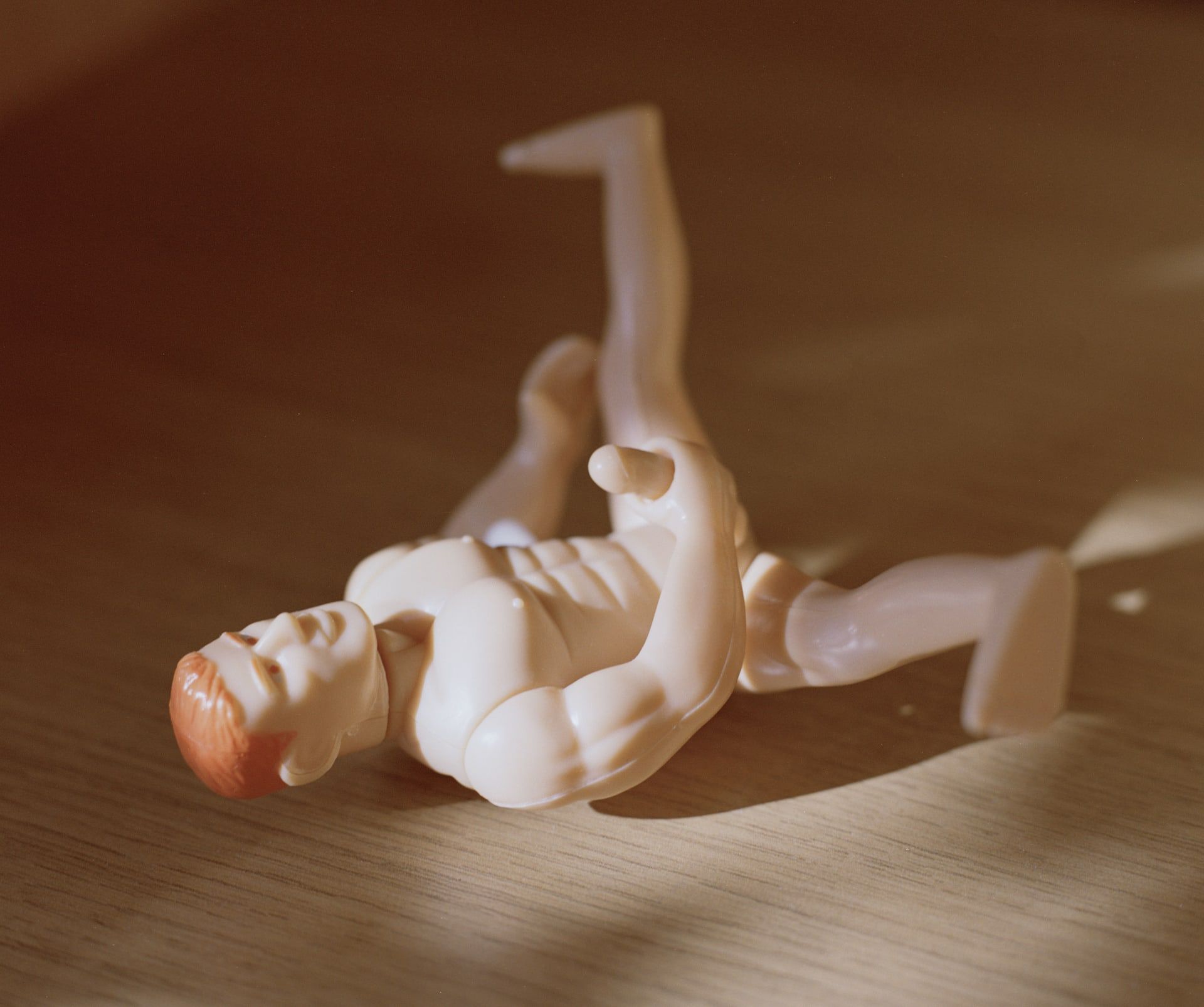 A photo of a small plastic muscle man toy holding it's penis laid on it's back on a wooden table