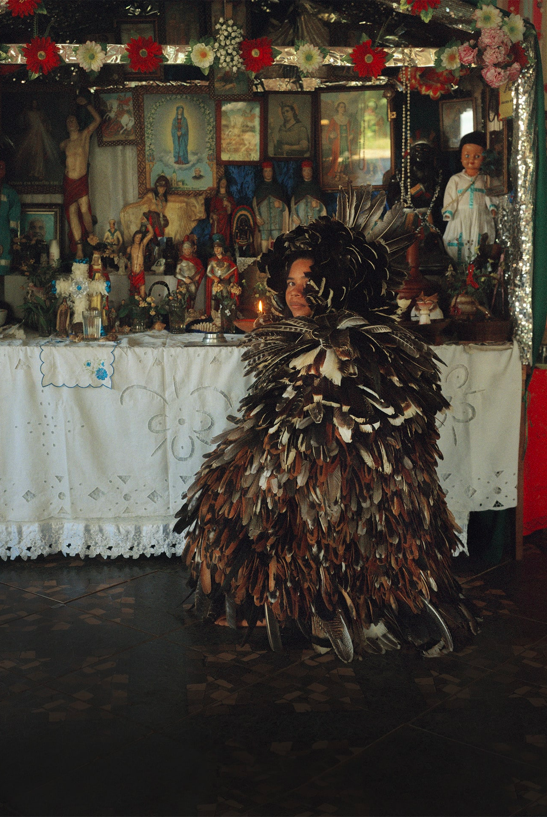 A photograph featuring a person completely dressed in feathers, crouching in front of a table decorated with model figures and paintings, turning their head to face the camera, by Fernanda Liberti. 