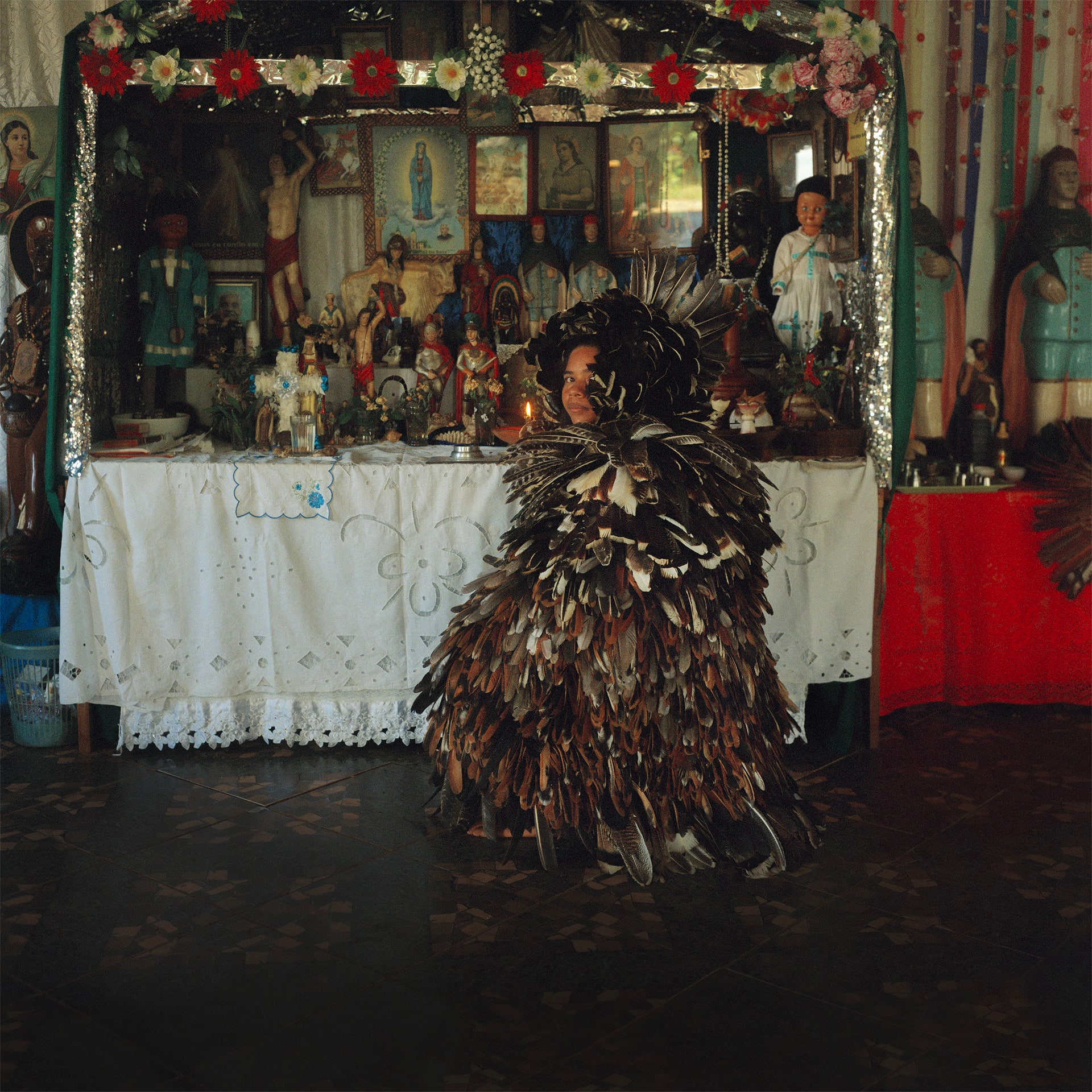 Photograph featuring a person completely dressed in feathers, crouching in front of a table decorated with model figures and paintings, turning their head to face the camera.