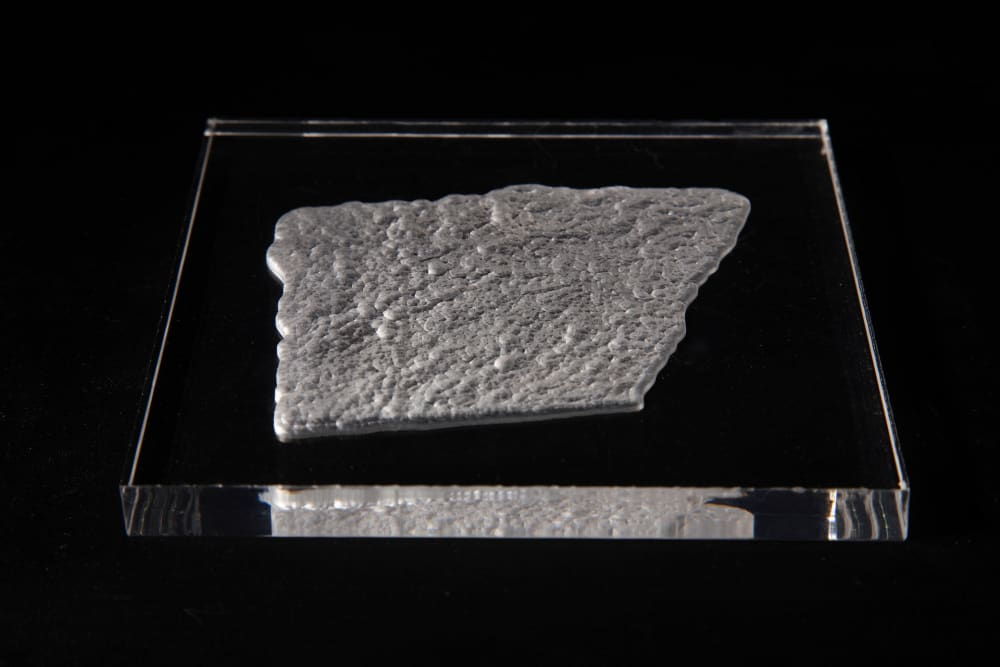 Black and white photograph of what appears to be an uneven square slice of rock or stone, with a pimply surface, contained in a thin, square block of clear glass, on a black background.