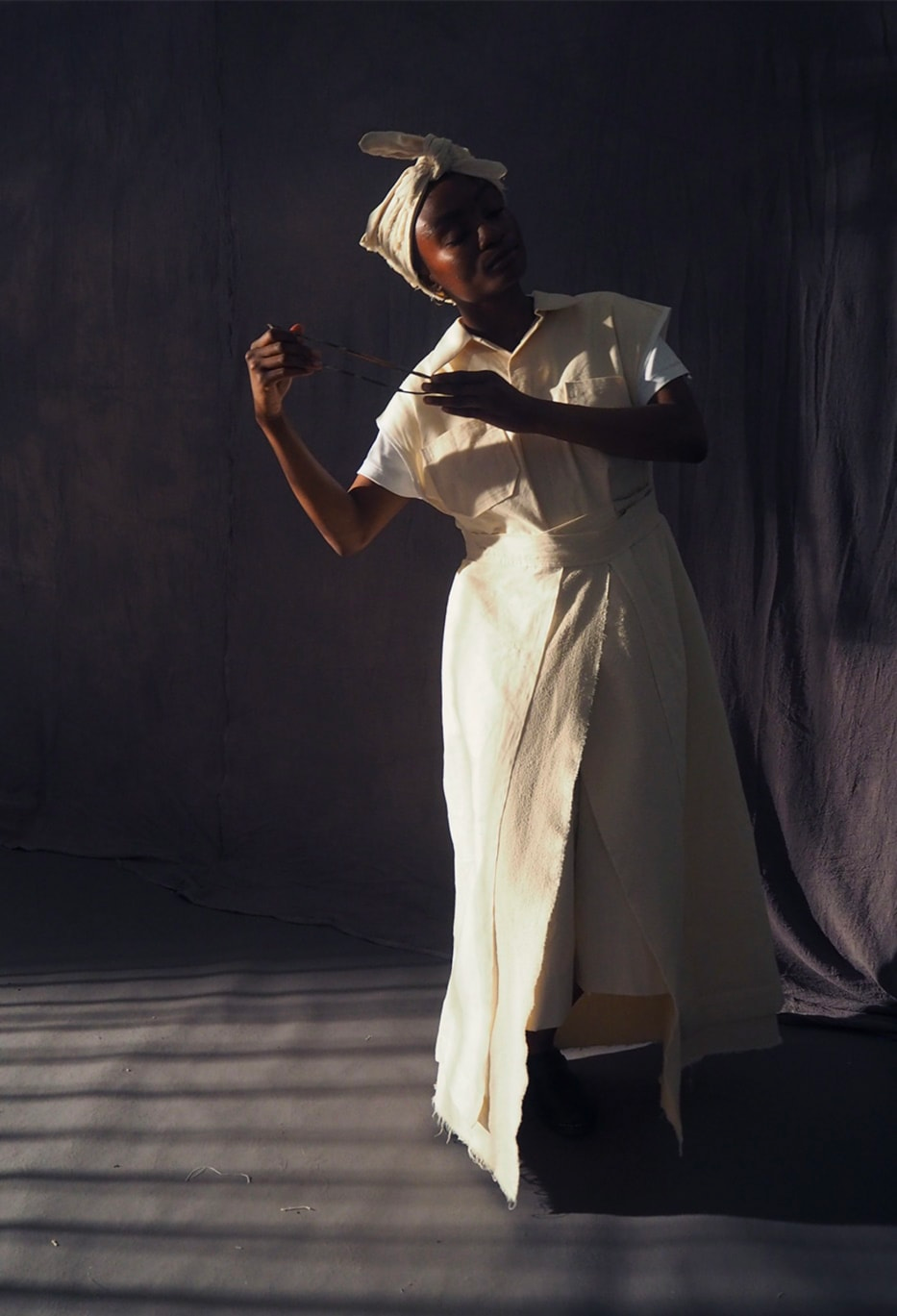 An image of a black woman in white-cream robes, partially in shadow, standing in a room with her eyes closed, holding an object, and leaning slightly to one side.