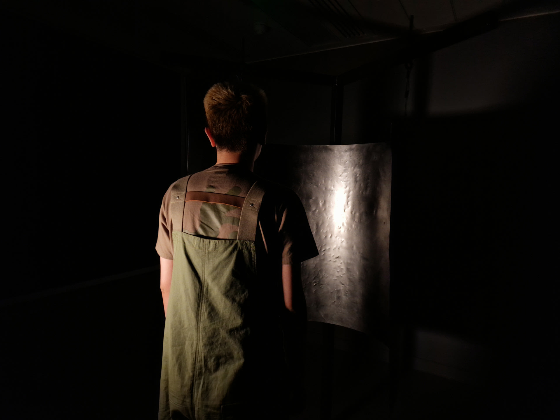 Photograph of a partially lit person, with their back to the camera, stood in front of a dented metal panel, in a completely dark background. 
