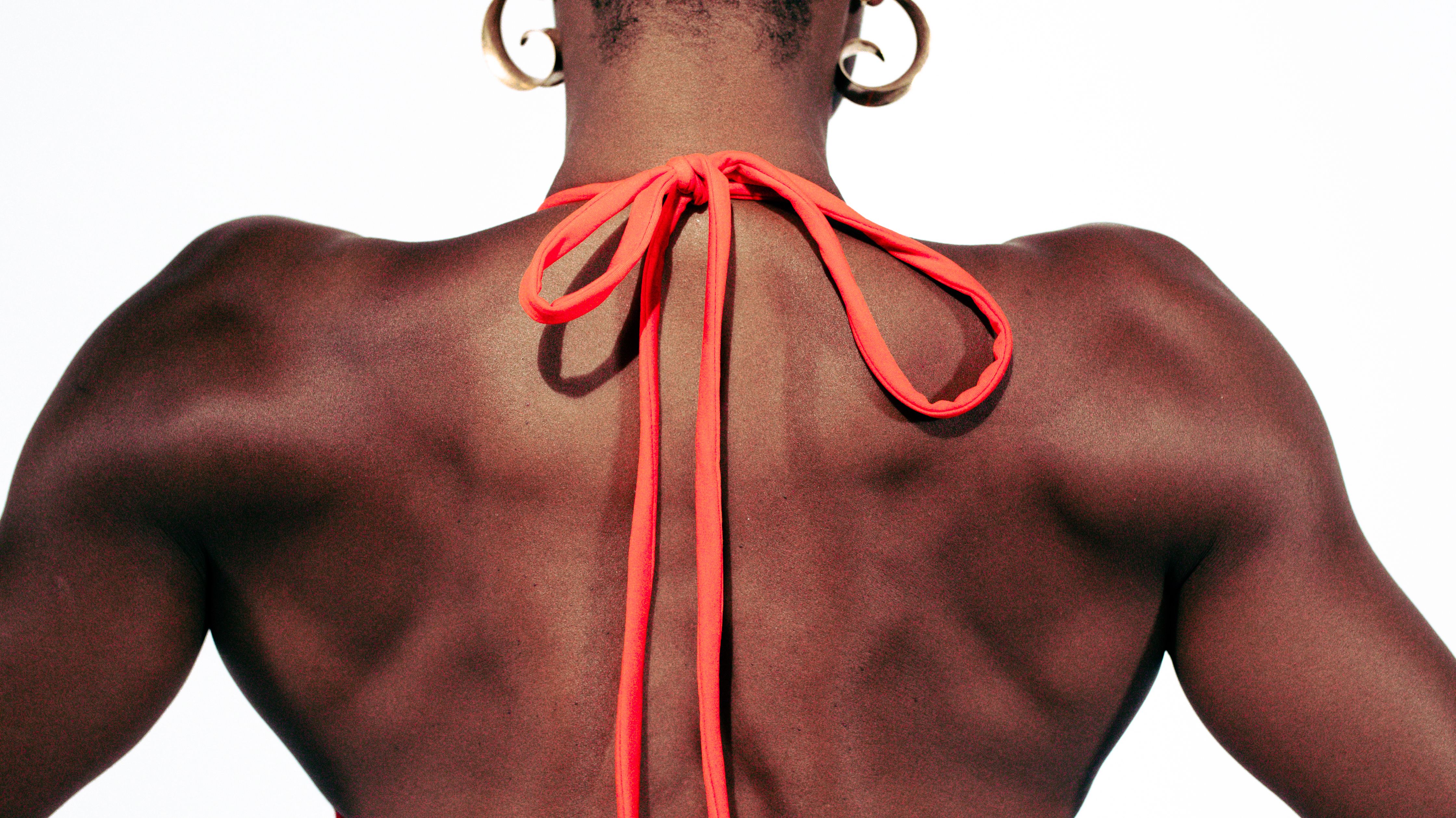 Photograph of the back of a woman from the waist up, wearing an orange red string bikini top, tied in bows at the neck and across the chest.