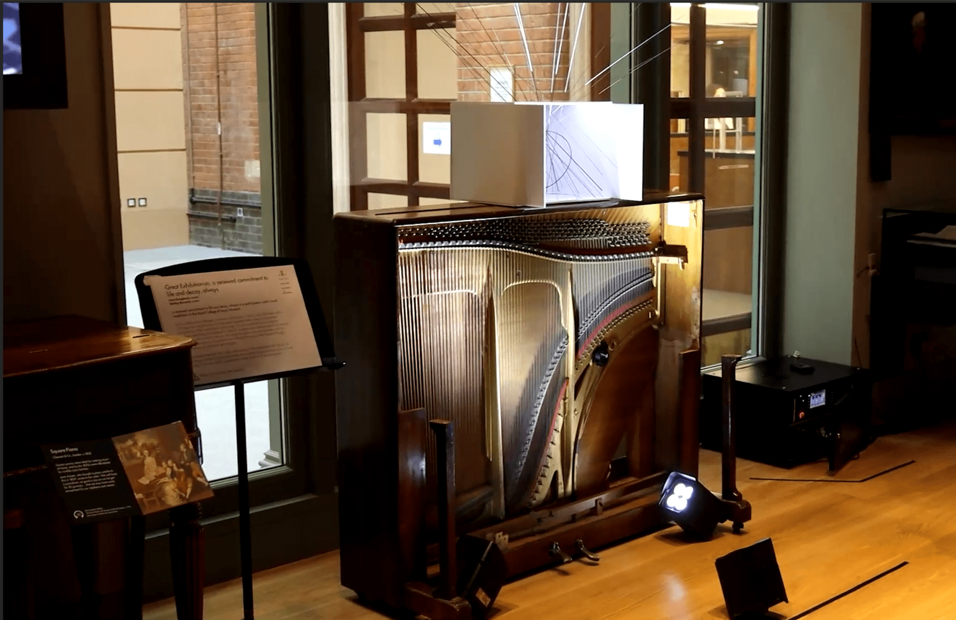 Photograph of objects, including a stand holding a printed page, and what looks like the back section of an upright piano, strings exposed, displayed in front of a window in a softly lit space.
