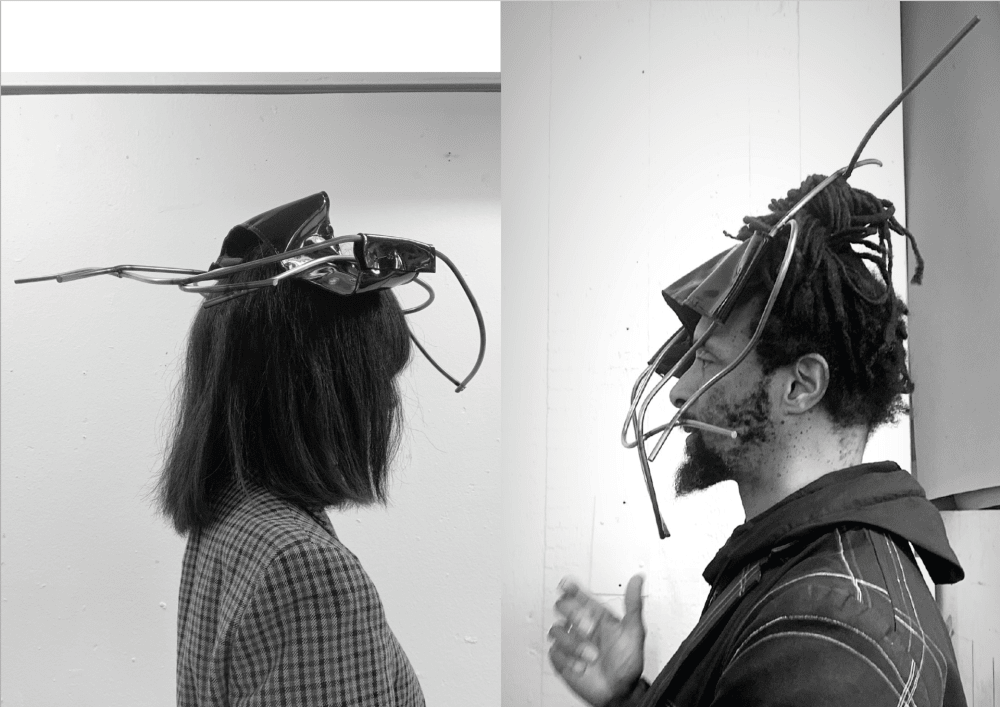 Black and white photograph of two people, from the shoulders up, in profile, facing each other, one turning their face away from the camera, and both wearing contraptions on their heads.