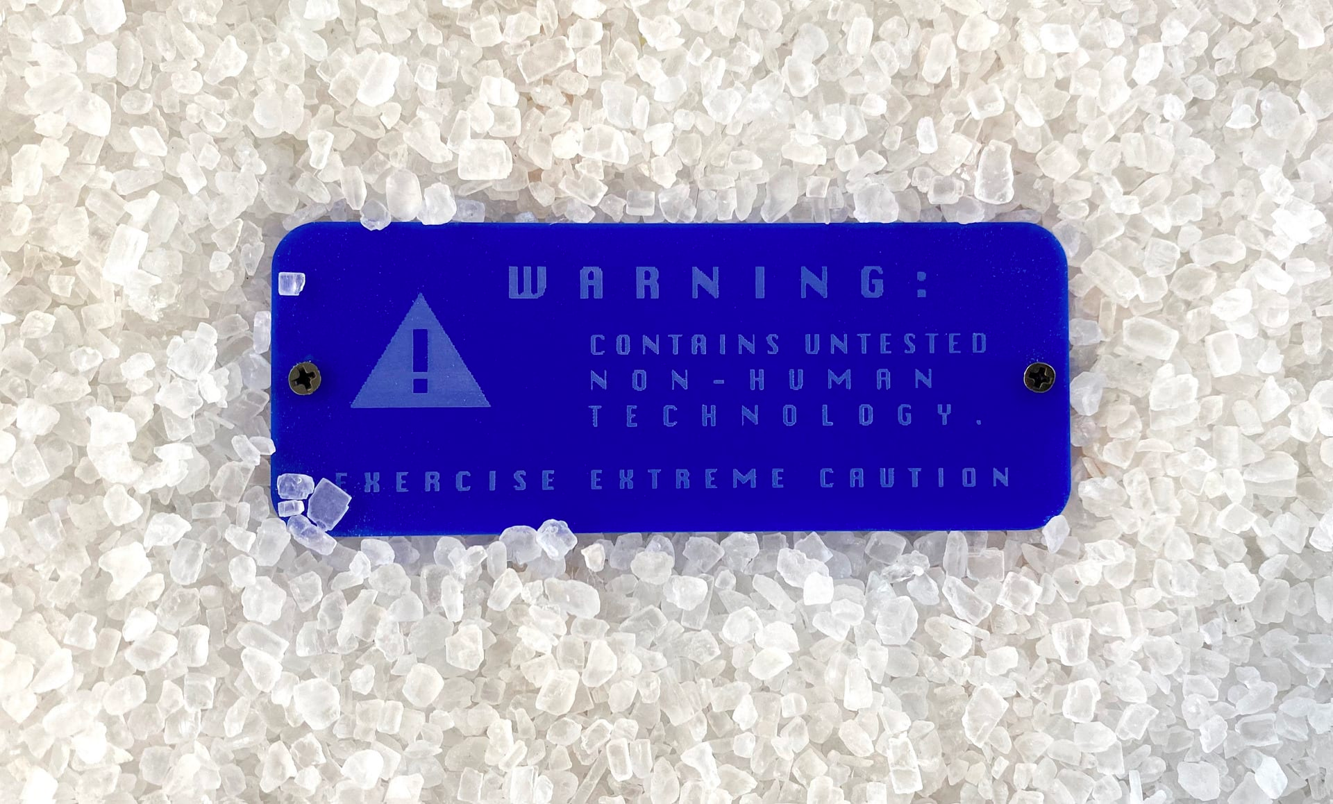 Image of a rectangular blue warning sign with two black screws saying, ‘contains untested non-human technology’, placed in the middle of what looks like salt crystals.