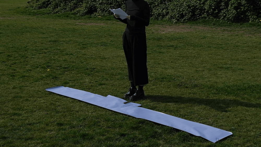 Photograph of a person from the neck down, dressed all in black, holding a sheet of white paper in front of them, and stood on grass with a long white strip or paper of fabric laid out in front of them.