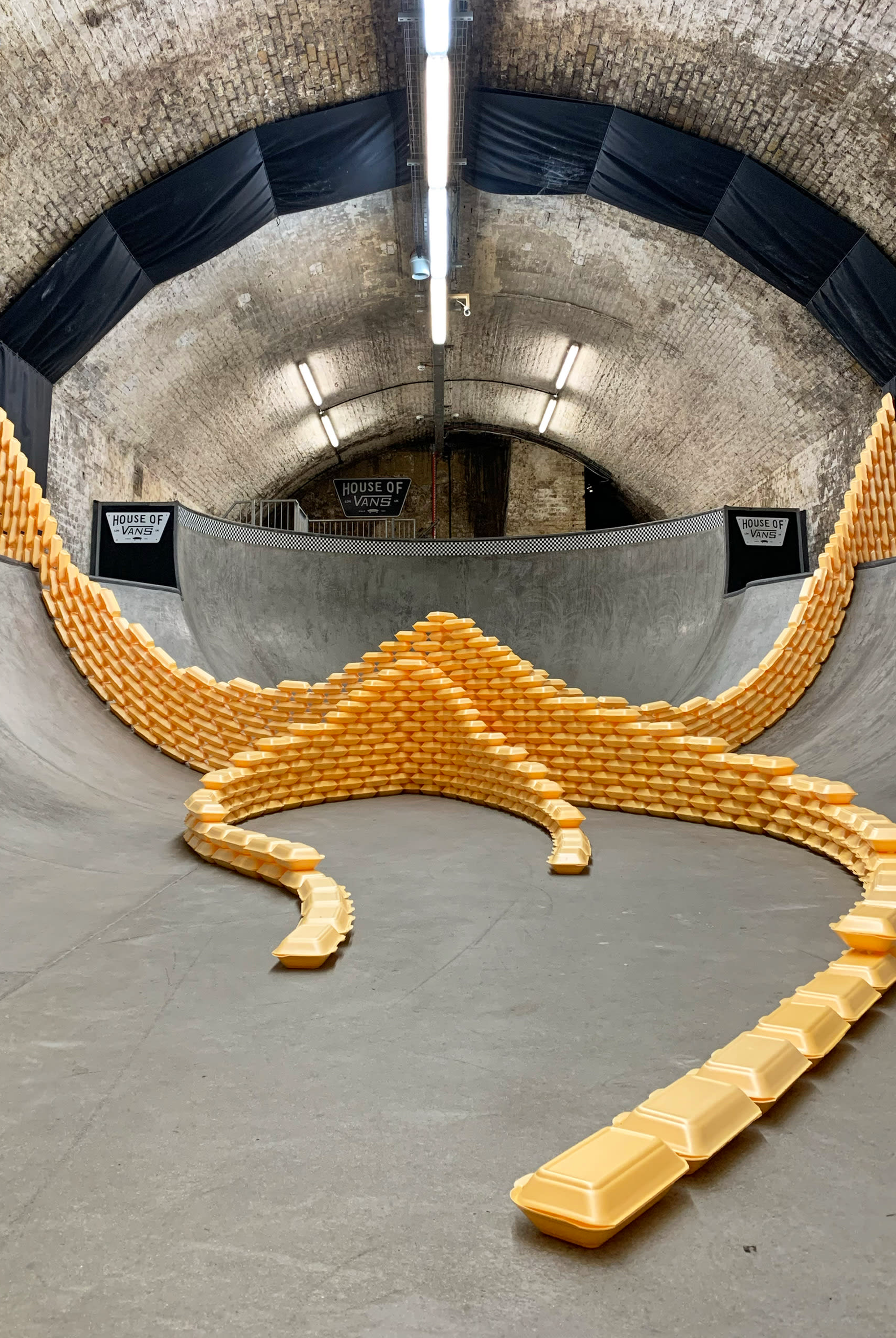  A skate-park inside a tunnel. Sweeping, curved, wall-like structures, composed of yellow polystyrene takeaway boxes are at the centre of the image. By Jacob Talkowski.