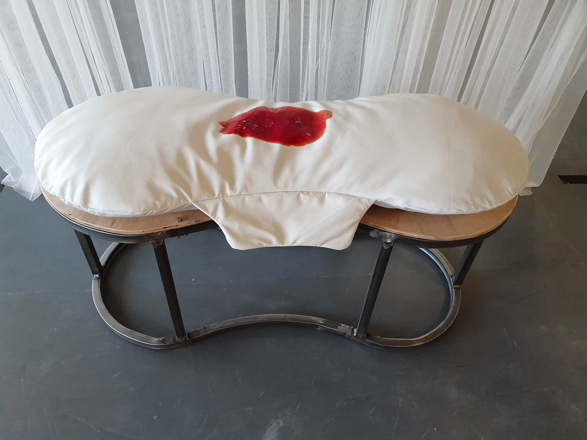 Photograph taken from above and in front of what looks like a curved, stool seat with a metal base, with a cream cushion shaped like a sanitary towel, and stained with red in the middle on top.