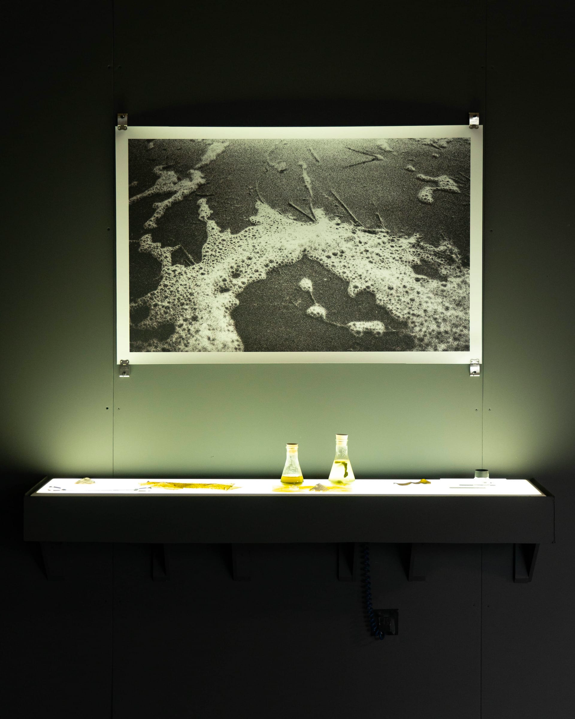 Image of what looks like a black and white photograph of sea foam on sand, hung on a wall above an illuminated shelf, which holds a couple of glass vase-like objects.