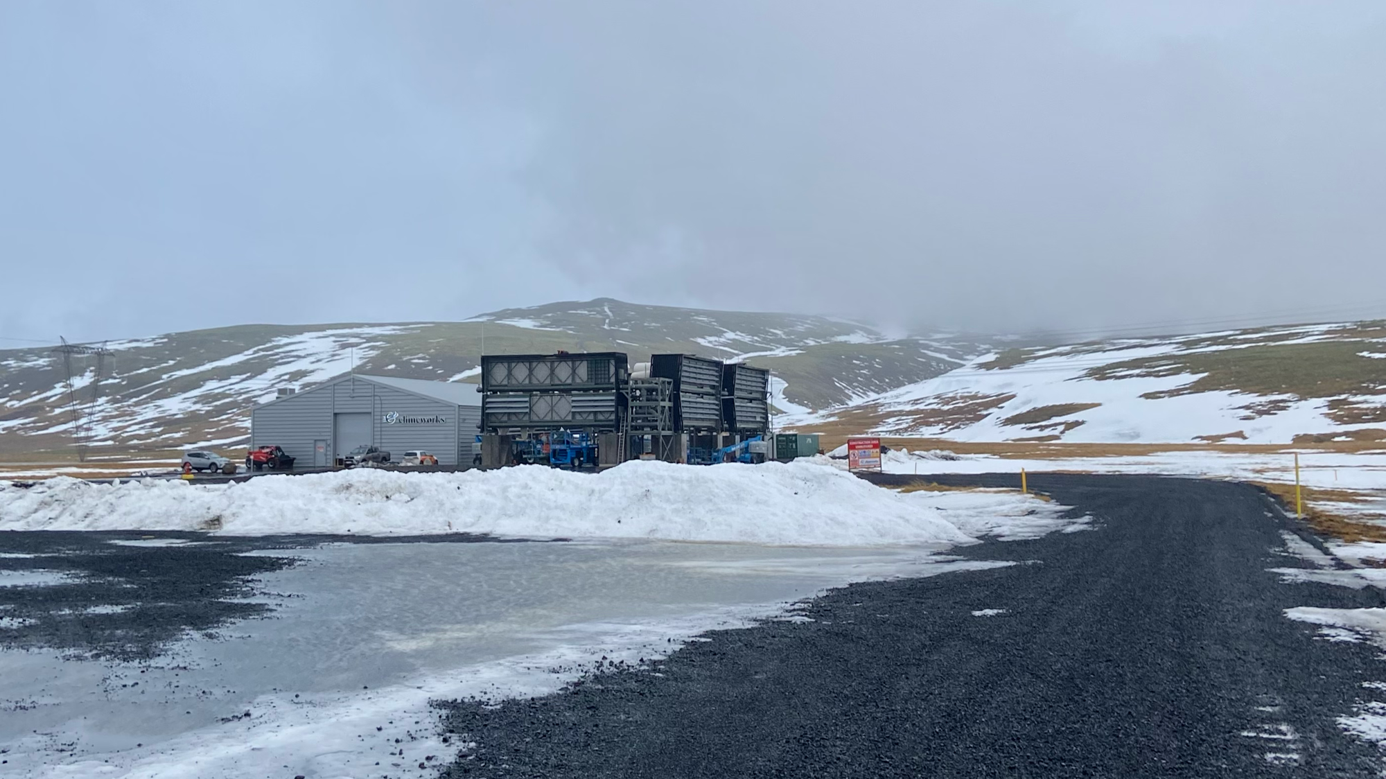 Photograph of a black unmade road leading to a large industrial-looking shed and equipment, with a few vehicles parked outside, in a deserted, partially snowy landscape with hills in the background.