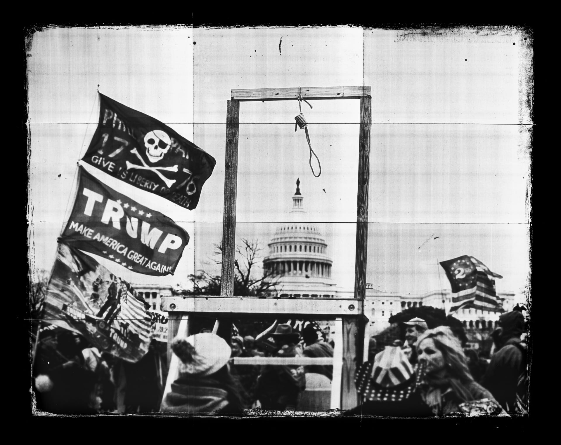 Image which features a black and white photograph of a crowd, with political banners, and a wooden gallows with a rope noose, framing the dome of the Capitol building in the distance. Flags visible to the left of the image show a skull and crossbones and read "Trump Make America Great Again"