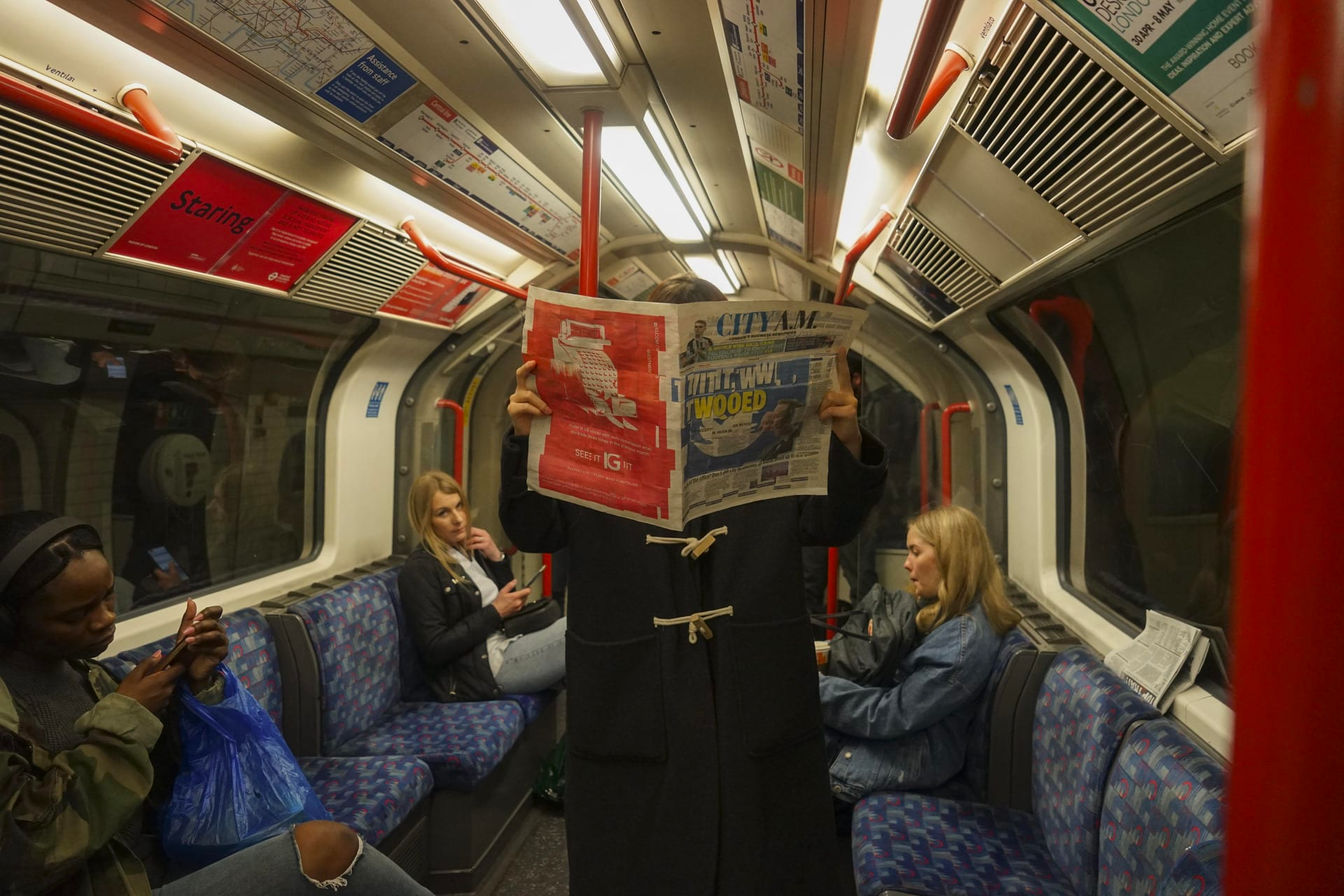 Photograph of a Central Line underground train carriage, with some seated passengers, and a passenger standing, facing the camera, holding an open newspaper, which covers their face.