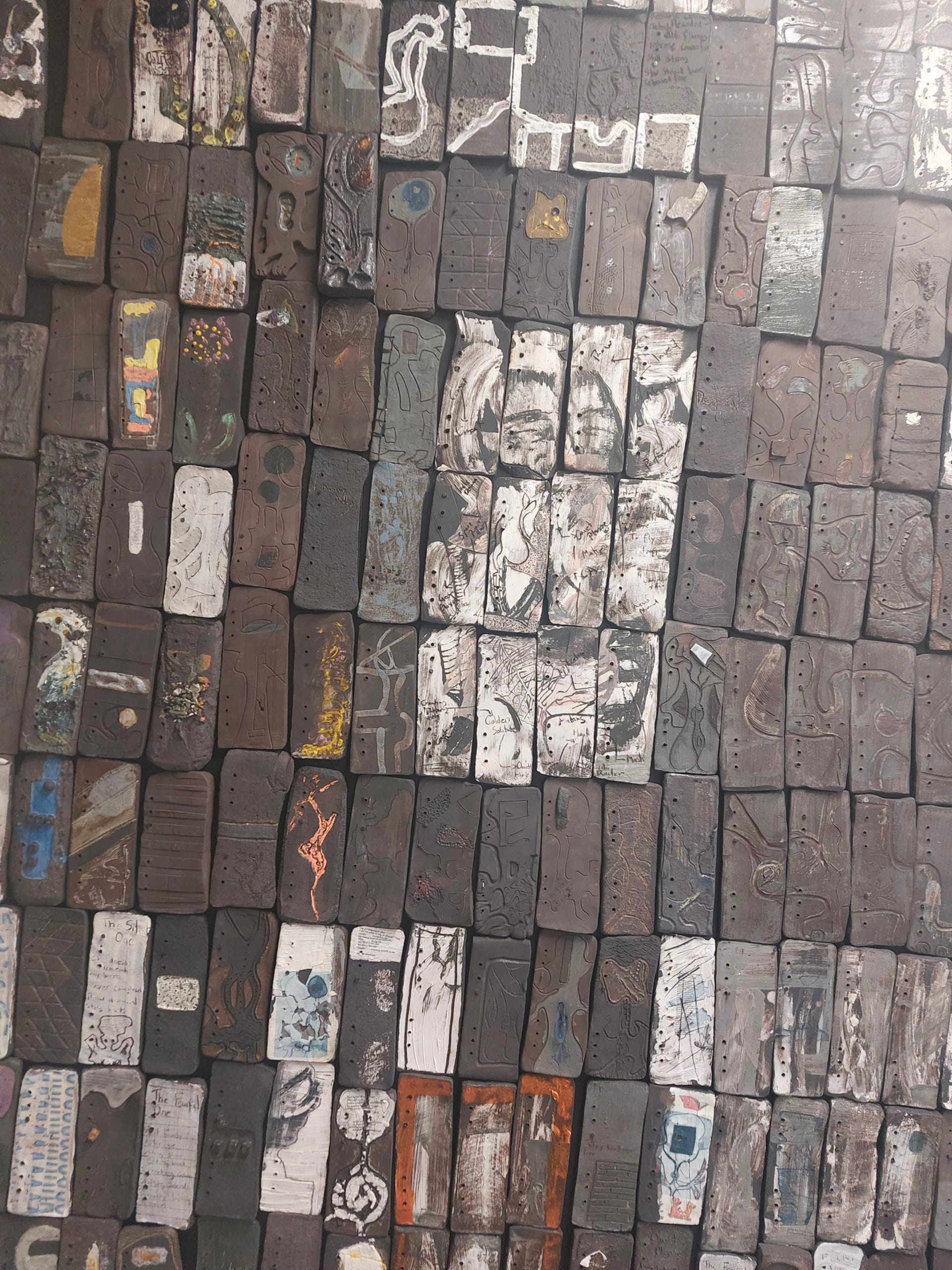 Image of many slightly wonky, brown rectangular bricks, all with different images carved, drawn or painted on them, laid together like a wall or paving.