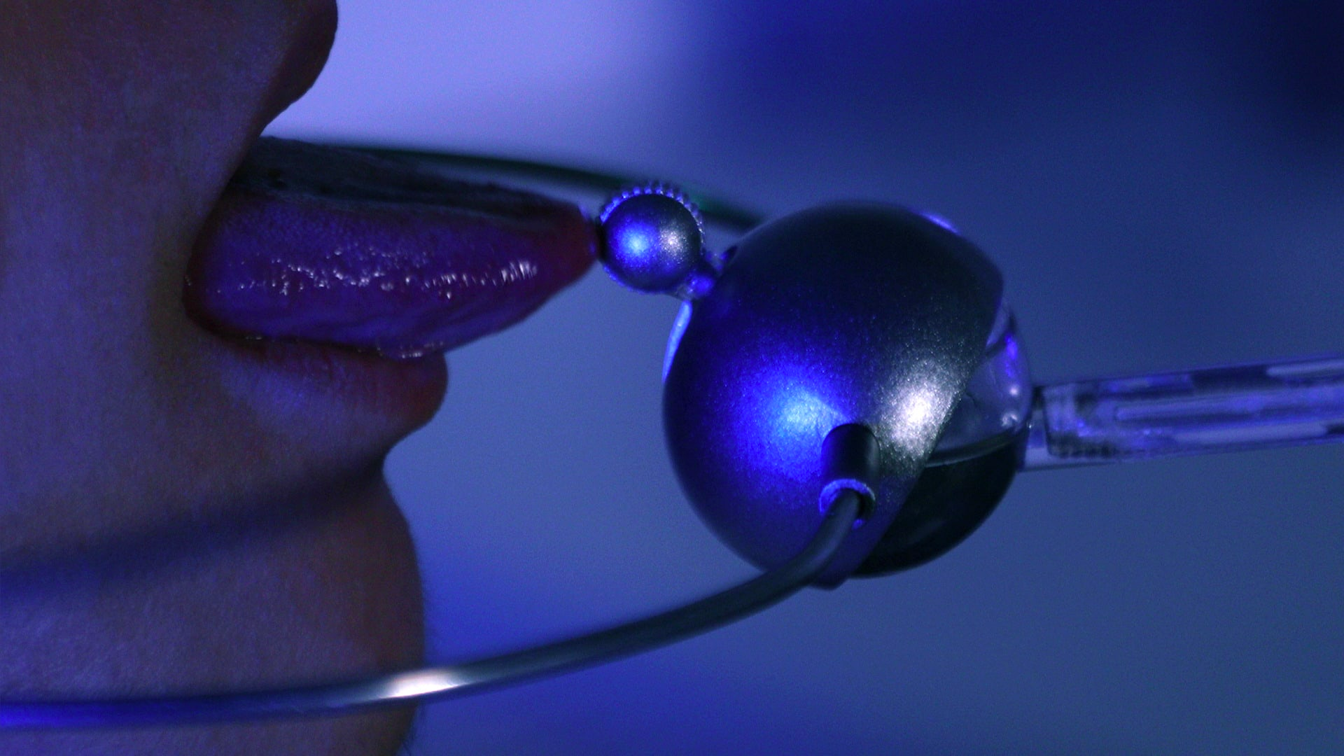 A blue-tinted photograph of a close-up mouth in profile, with the tongue protruding and its tip touching a small metal ball attached to some sort of device which includes a larger spherical object.