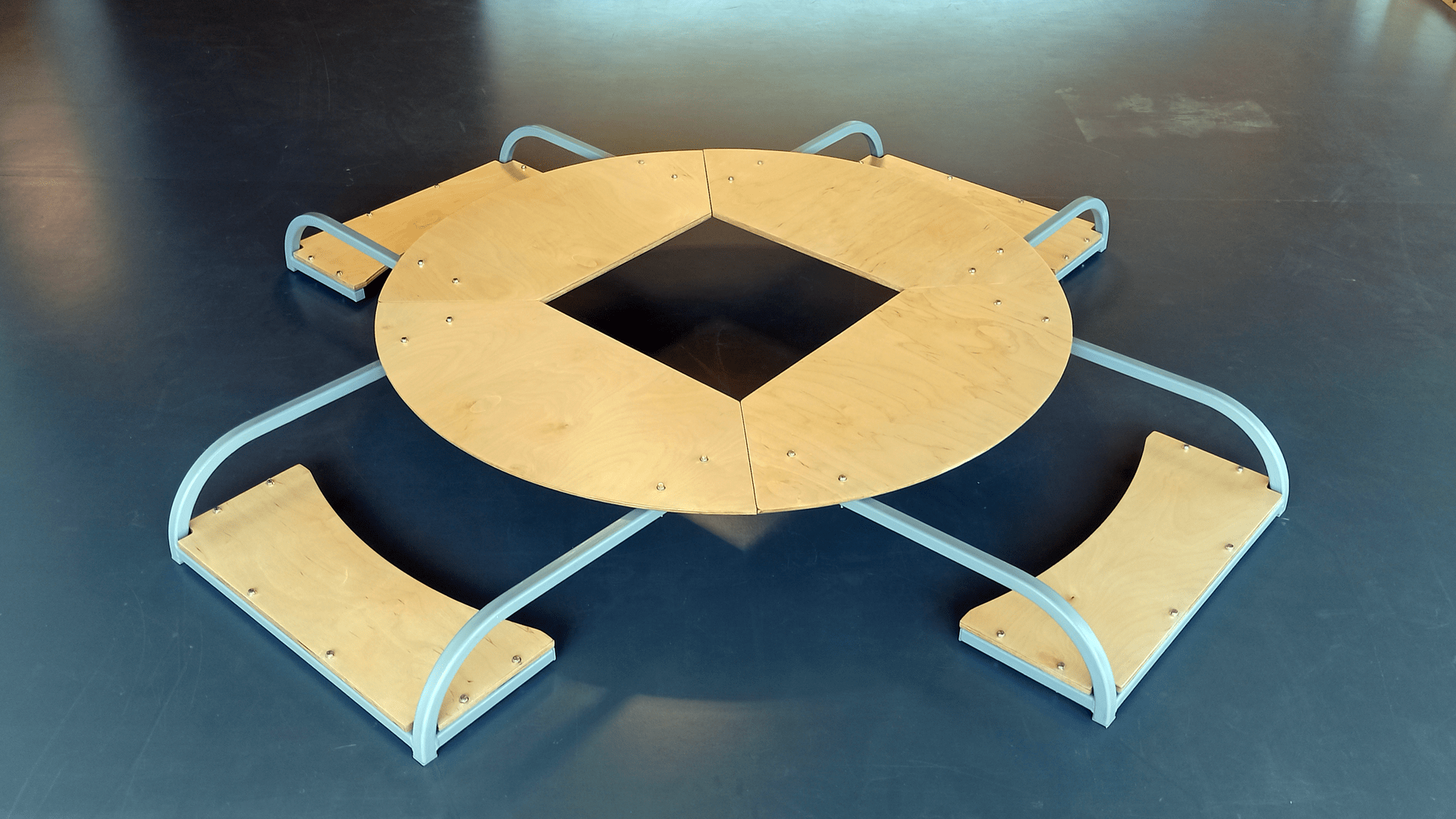 Image of what looks like a round wooden table, with a square cut out of the middle, with four wooden seats, which sit flat on the ground, attached to the table with curved metal bars.