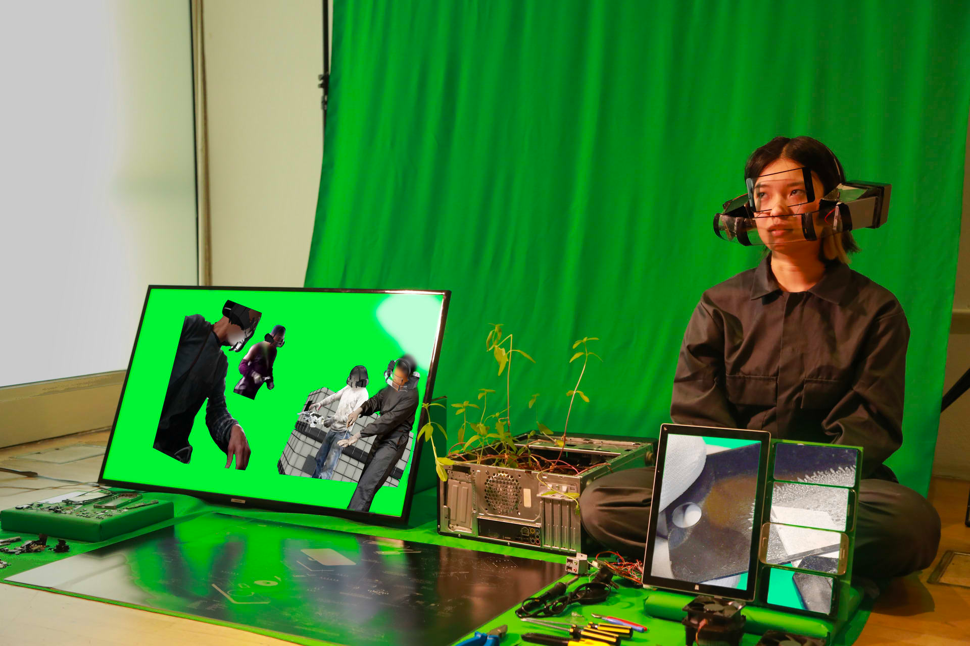 Photograph of a woman sat on the floor in front of a green curtain, next to an arranged collection of tools and objects with screens, wearing a contraption on her head which includes a clear screen over her eyes.
