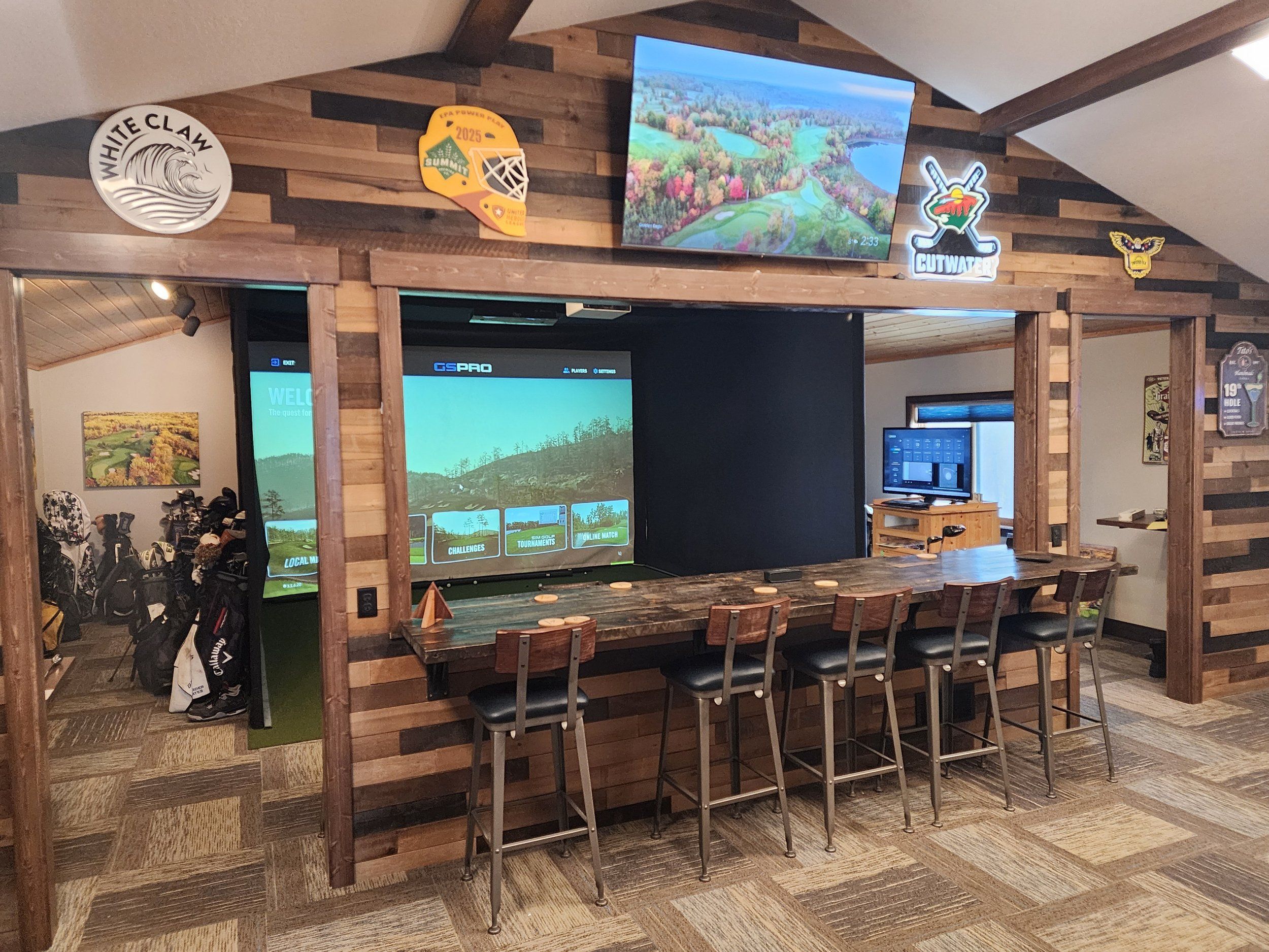 Simulator Lounge with a wood-paneled bar and stools facing the GSPro screen, plus TVs and a rack of golf bags