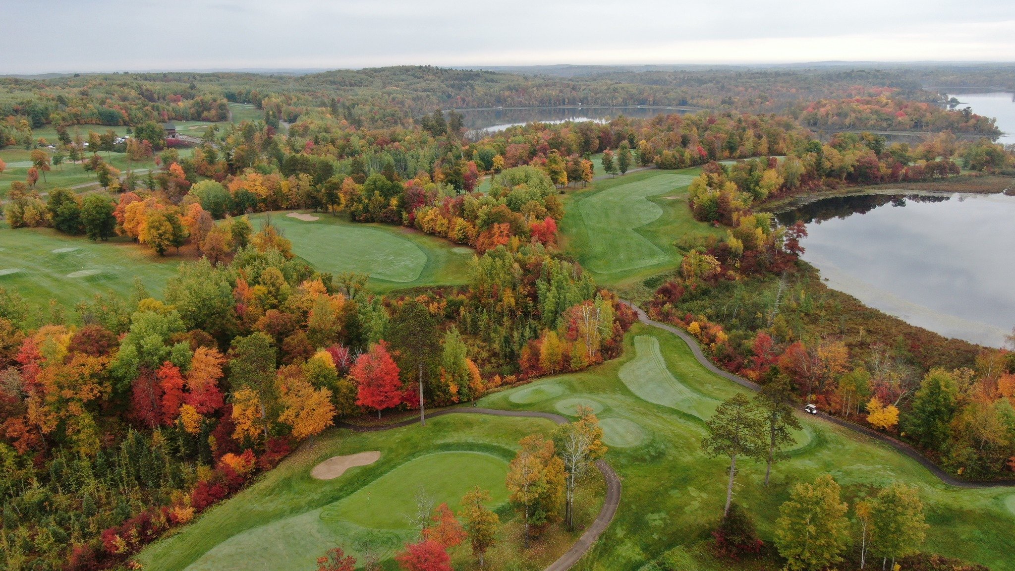 Aerial view of Golden Eagle Golf Club's fairways and greens surrounded by red, orange, and gold fall foliage, with lakes in the distance